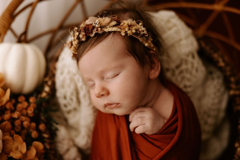 Sleeping newborn in autumn-themed setting with pumpkins and floral crown in warm earthy tones.
