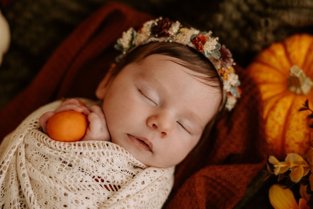 Sleeping baby wrapped in knit blanket with autumn pumpkin decor and floral headband in warm fall setting.
