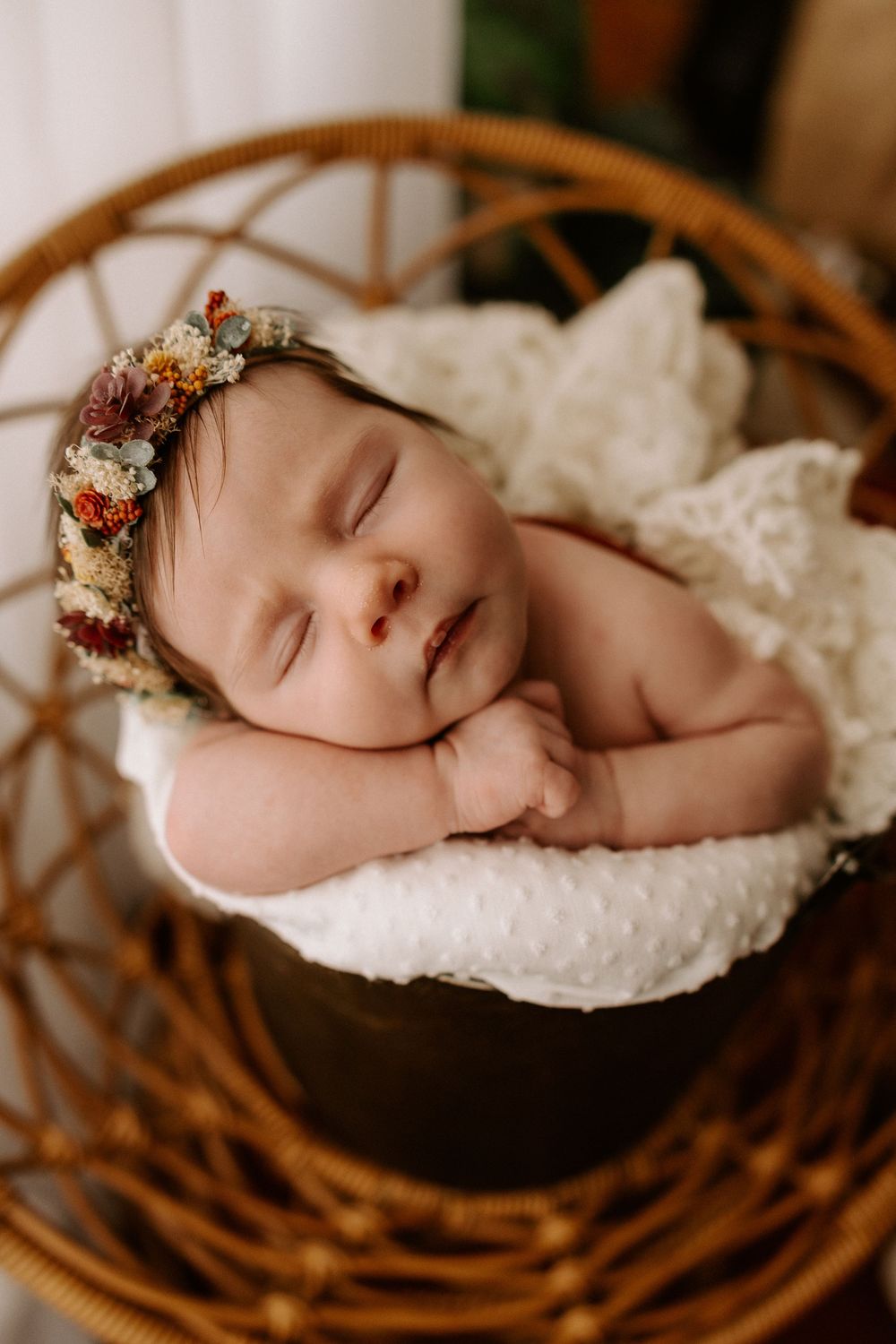Newborn baby sleeping peacefully in a rustic woven basket with white bedding and delicate floral headpiece.