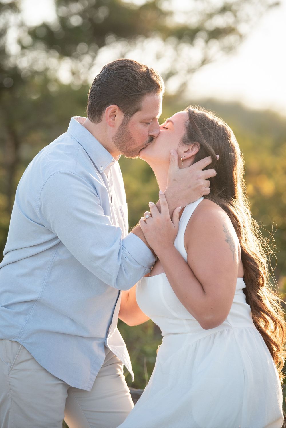 Romantic couple sharing tender kisses in golden sunlight wearing white dress and light blue shirt.