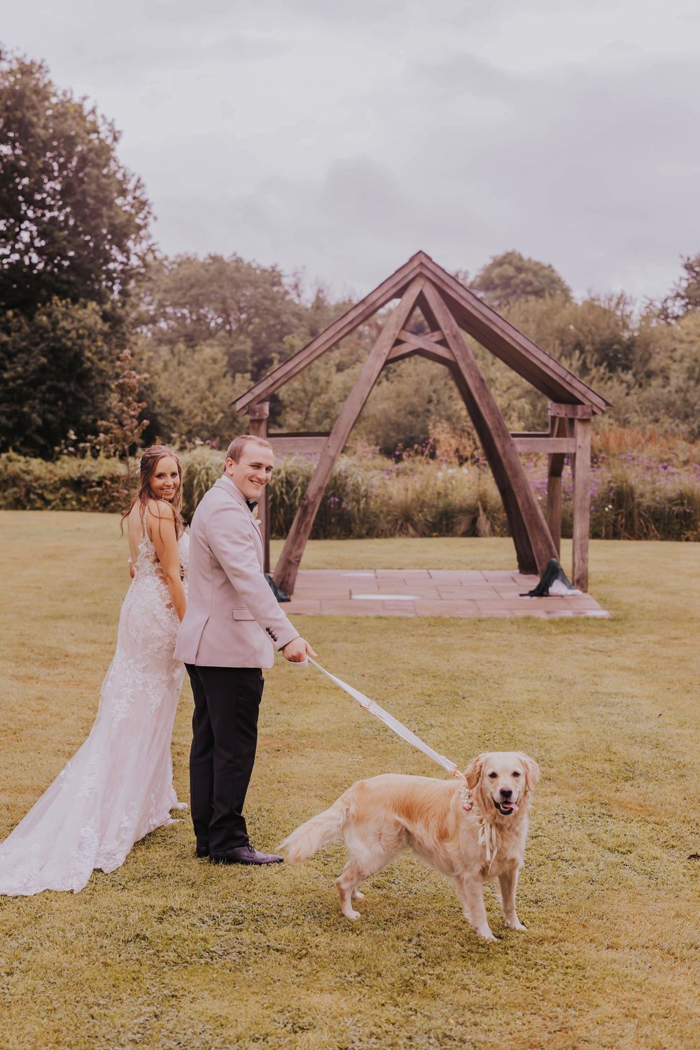 A couple in wedding attire walks their golden retriever through a grassy field with wooden structures in the background.