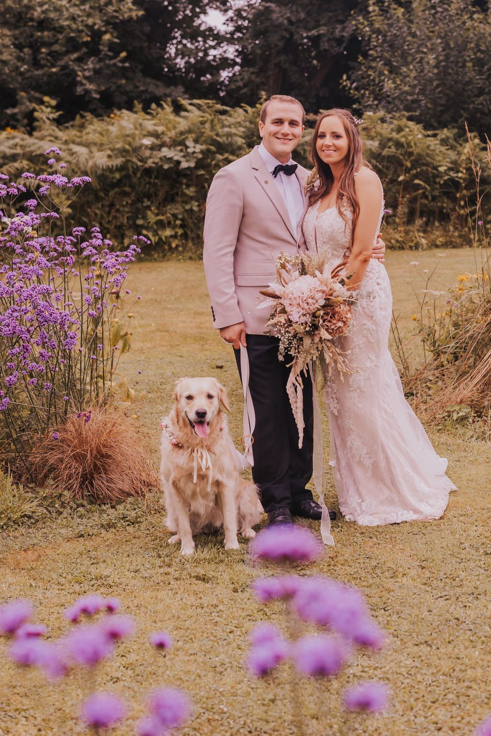 A couple poses with their golden retriever in a garden with purple wildflowers during a romantic outdoor photoshoot.