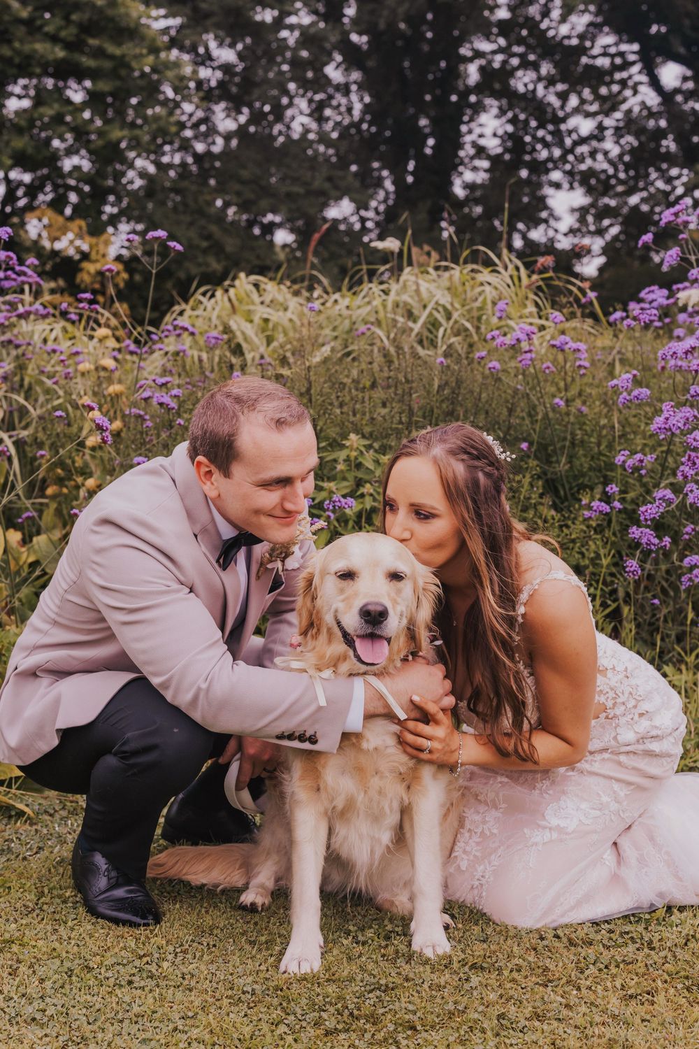 A couple in wedding attire shares a tender moment with their golden retriever in a flower-filled meadow.
