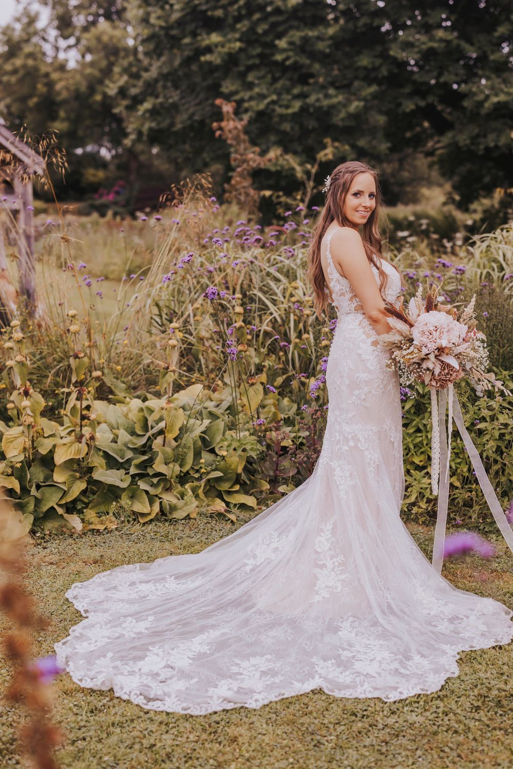 A bride in a flowing lace wedding gown with a long train poses in a garden setting during a summer photoshoot.