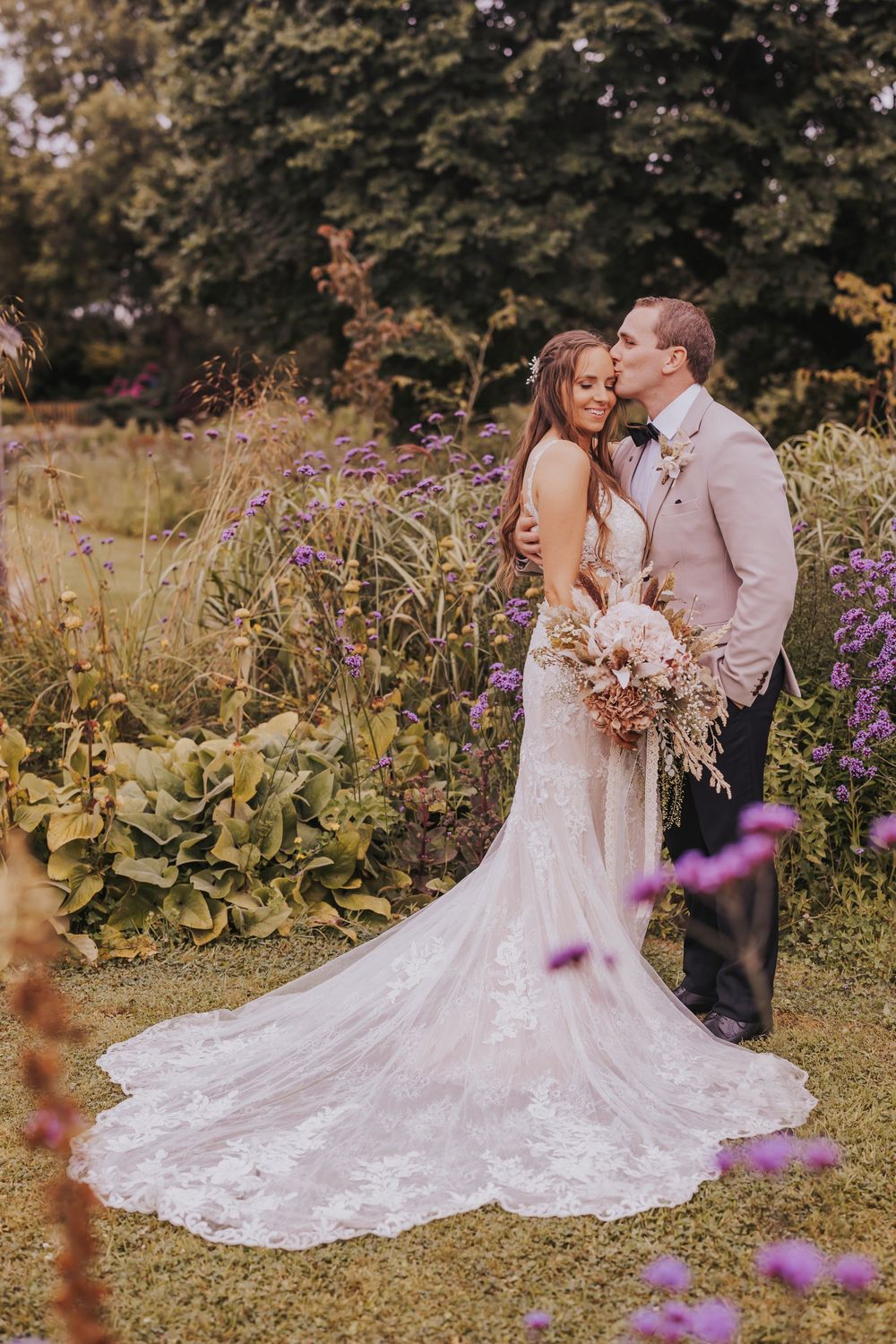 A romantic wedding photoshoot in a garden setting with purple wildflowers and flowing white lace dress.