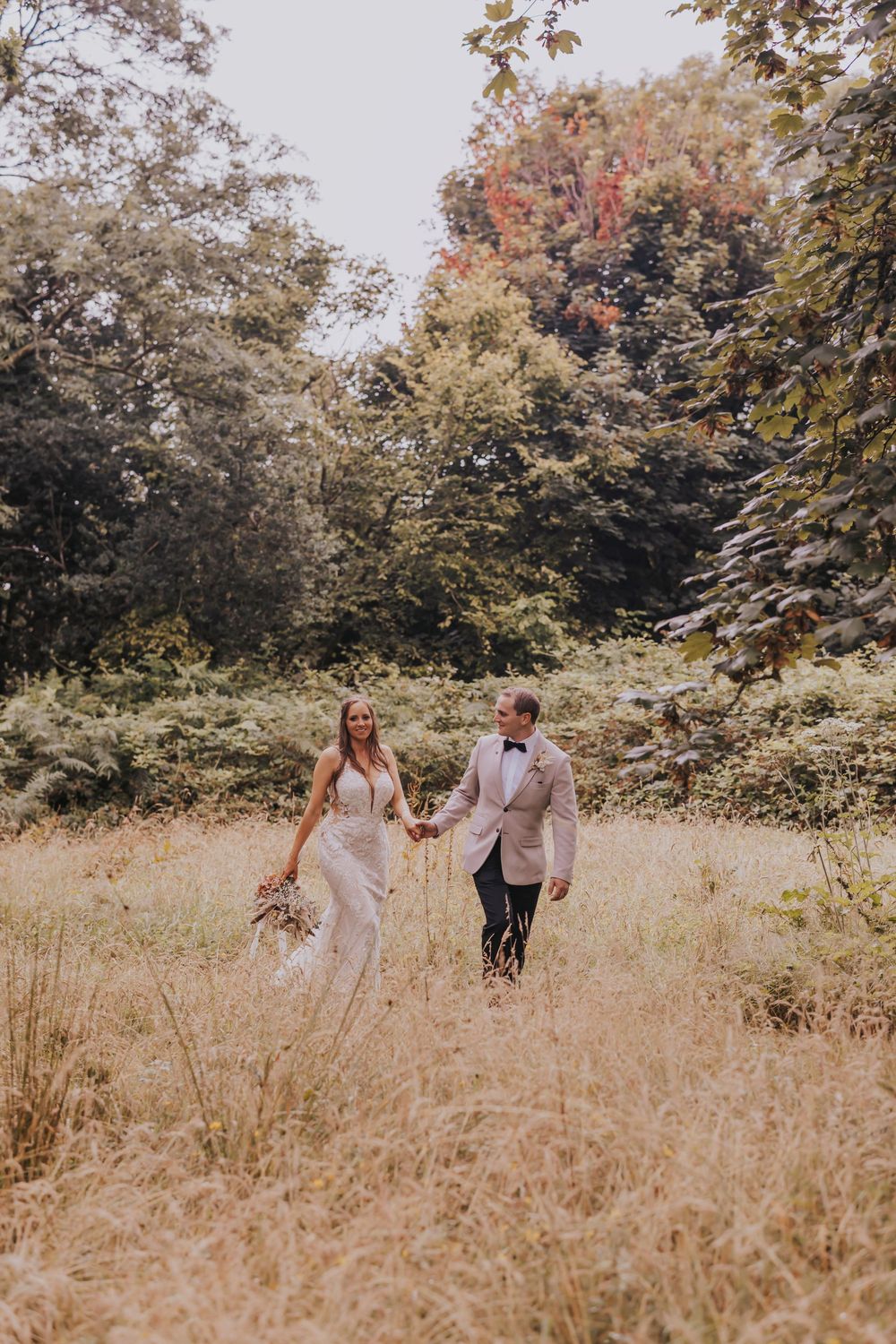 A couple walks through a sun-drenched meadow with tall golden grass and autumn trees in the background.