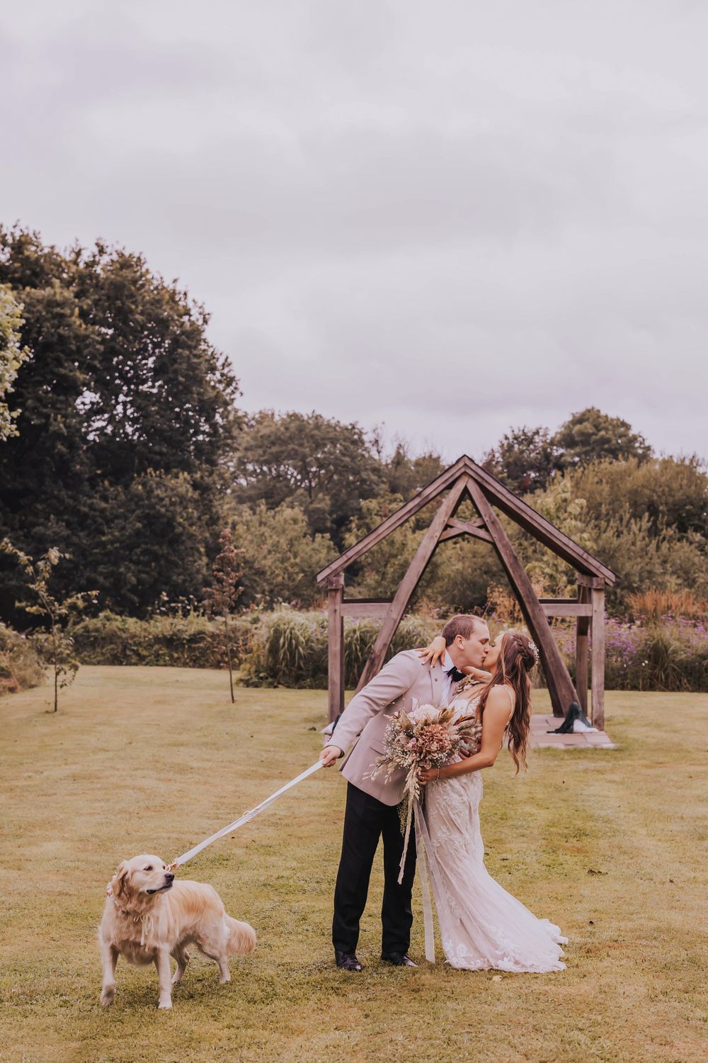 A couple shares a romantic moment under a wooden arch in a grassy field with their dog watching nearby.