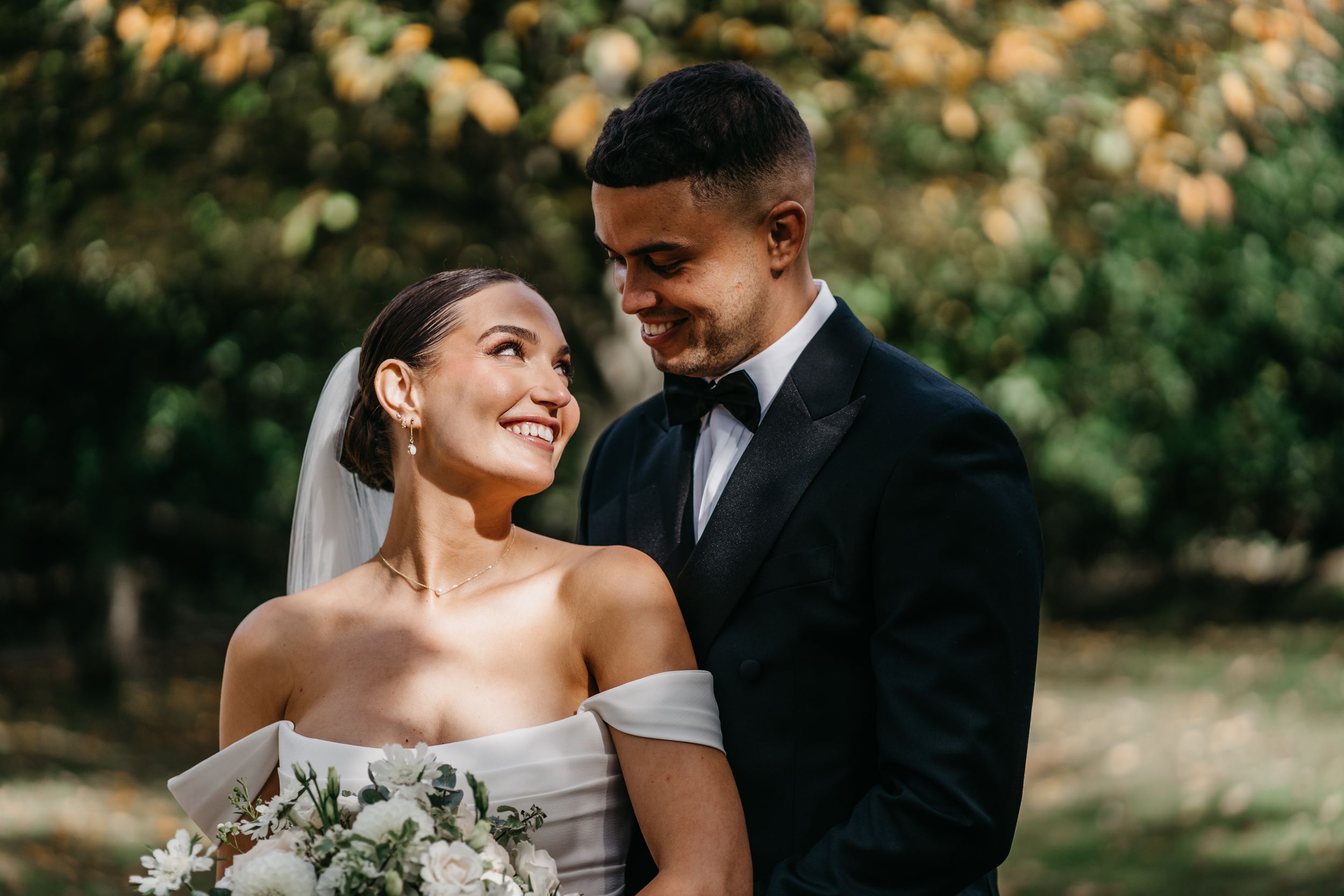 A romantic wedding couple embraces outdoors in a natural setting with soft lighting and greenery in the background.