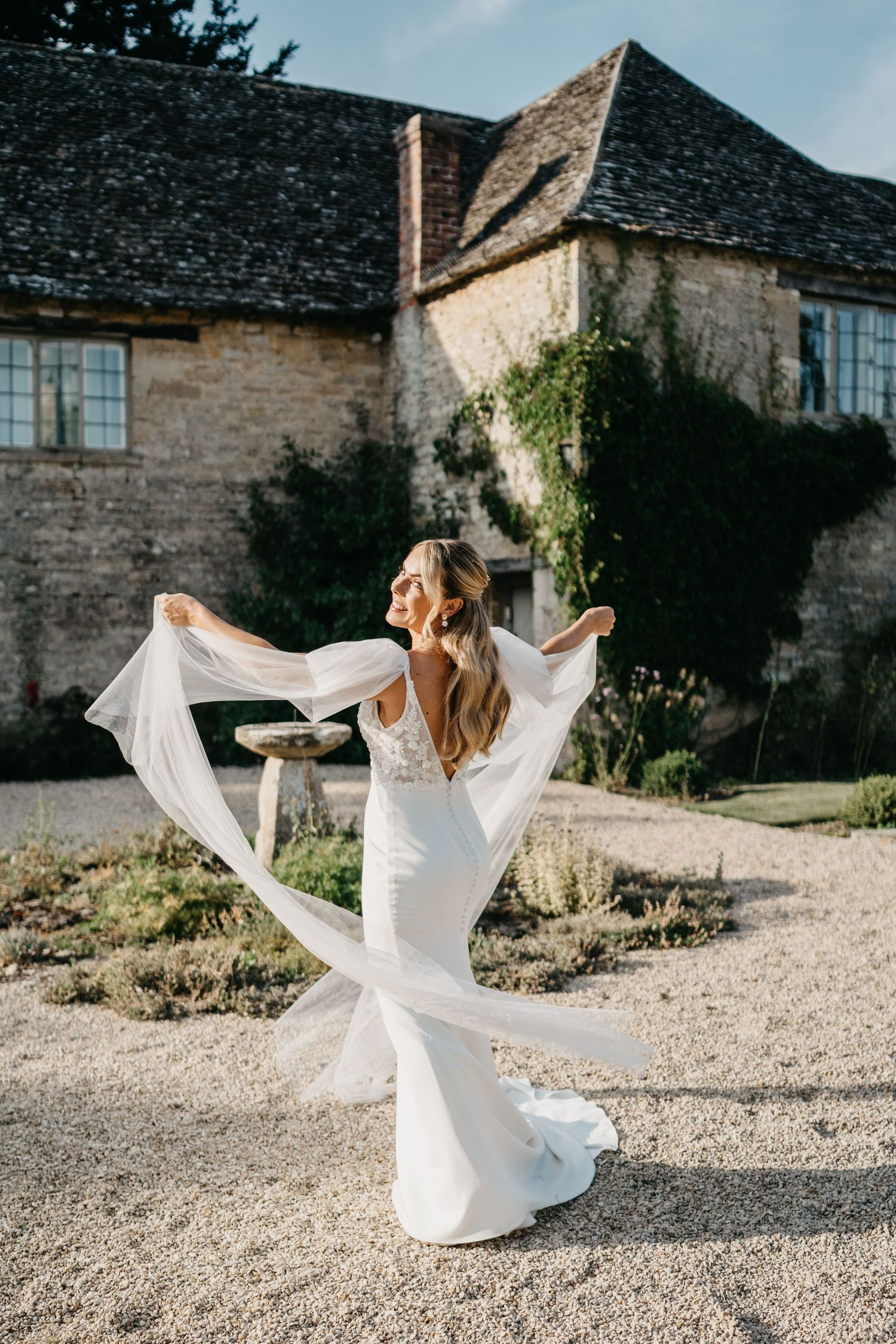 A figure in a flowing white wedding dress twirls gracefully on a gravel path beside a rustic stone building.