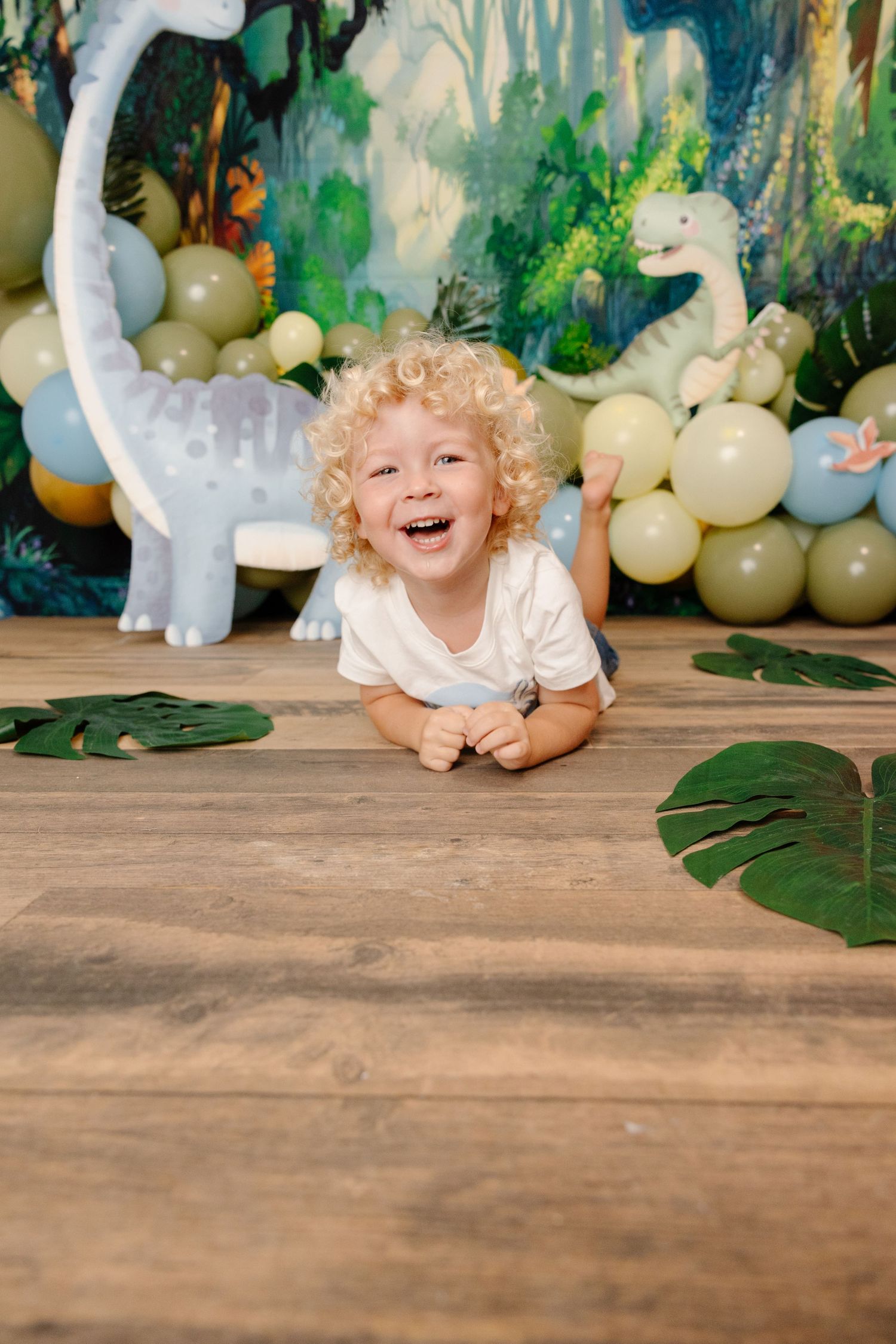 Child with curly hair lying on wooden floor with tropical themed balloon decorations and greenery in background.