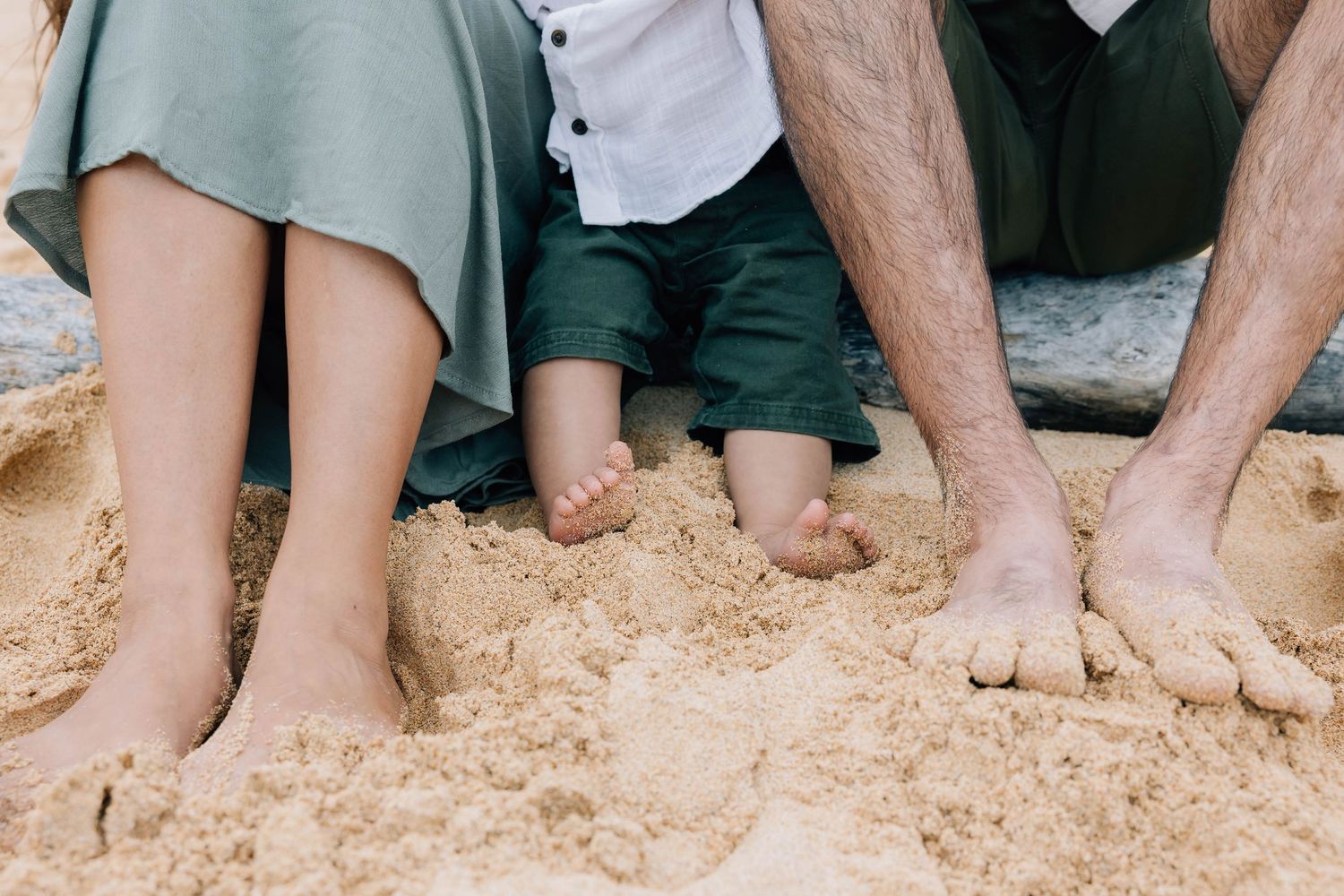 Barefoot family with baby sitting on sandy beach, close-up of feet in sand.