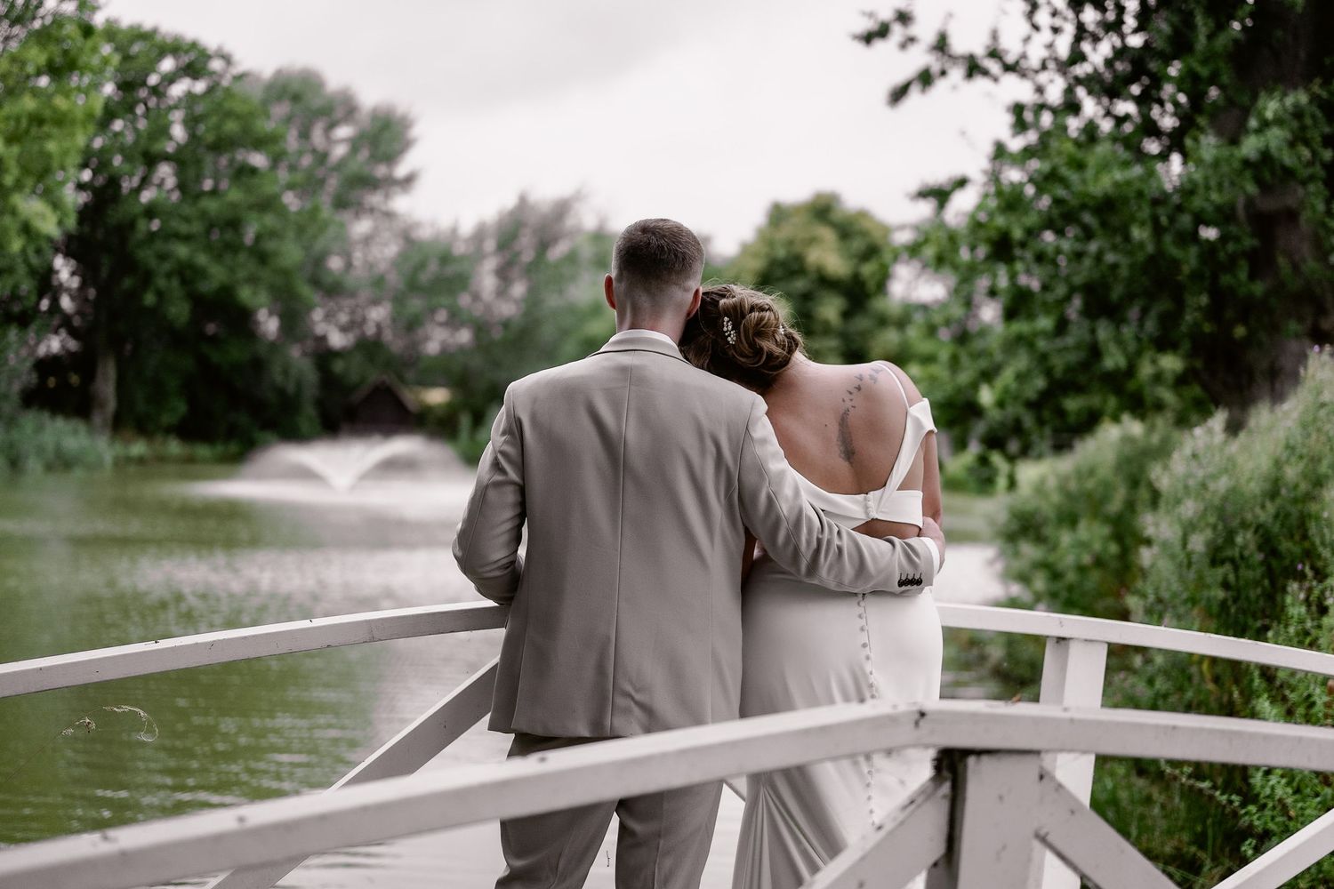 A couple in wedding attire stands close together on a white bridge overlooking a peaceful lake surrounded by trees.