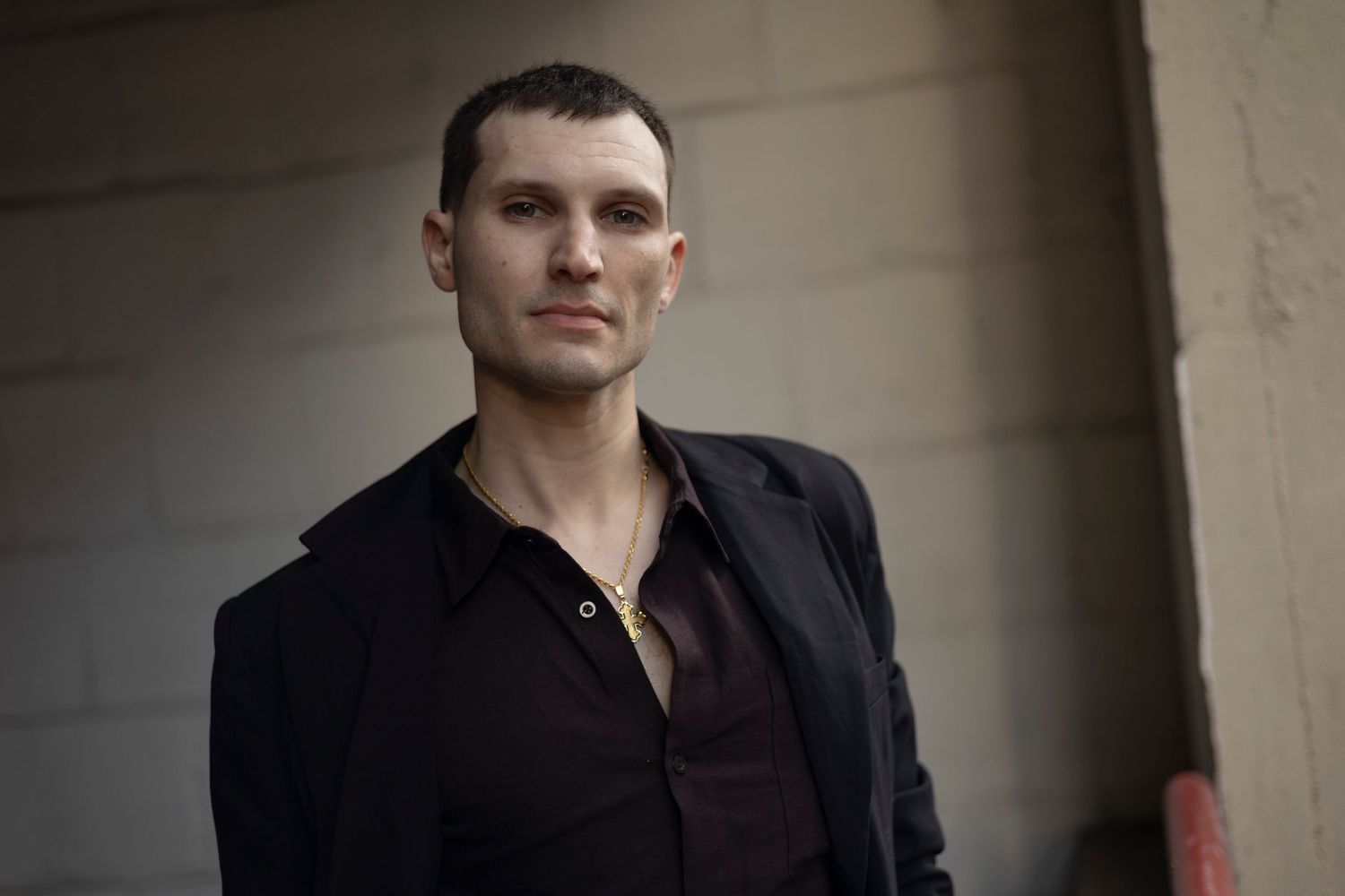 A person in a dark shirt poses against a white wall in dramatic lighting.
