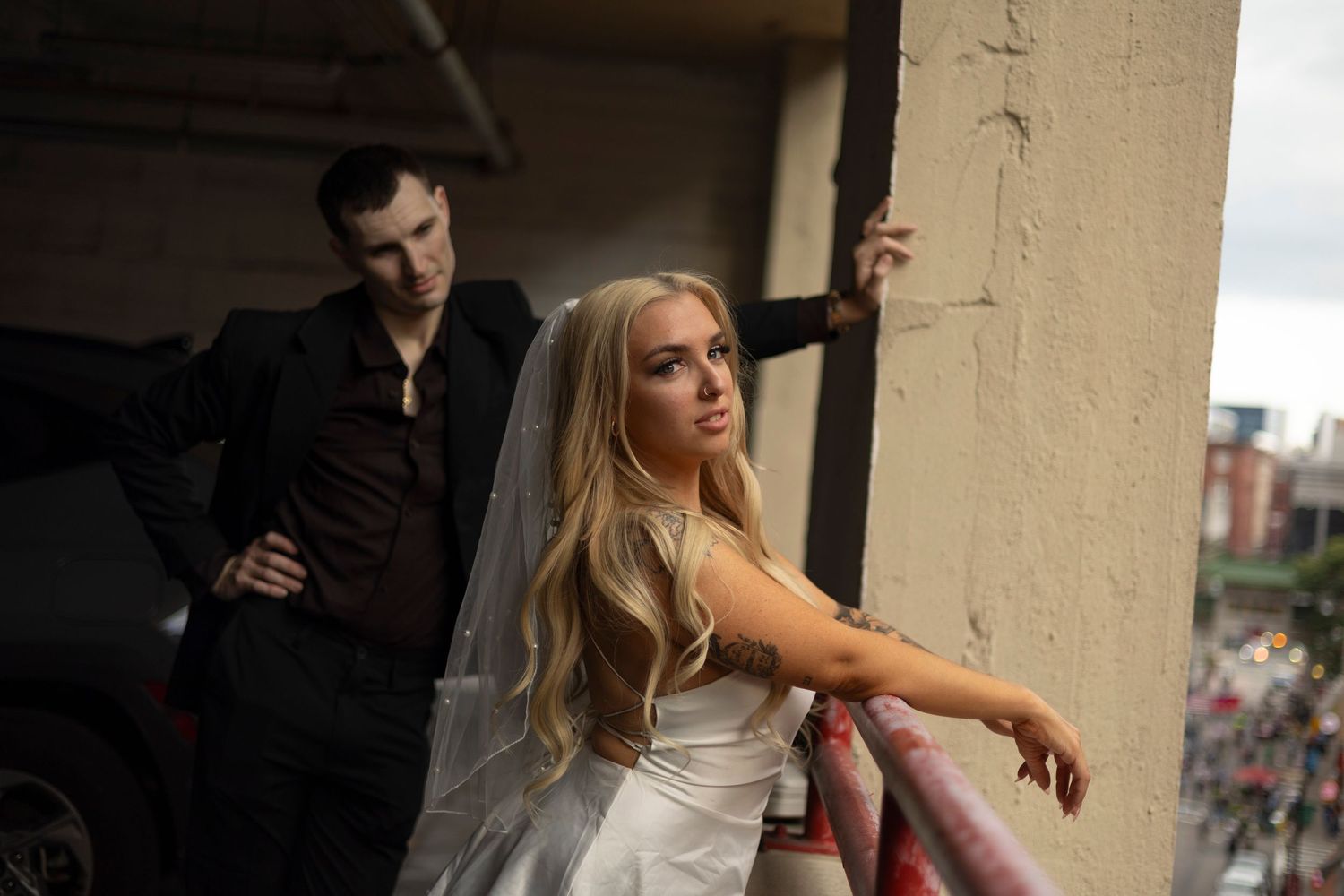 A bride in a white dress leans against a pillar while someone in dark clothing stands in the background.