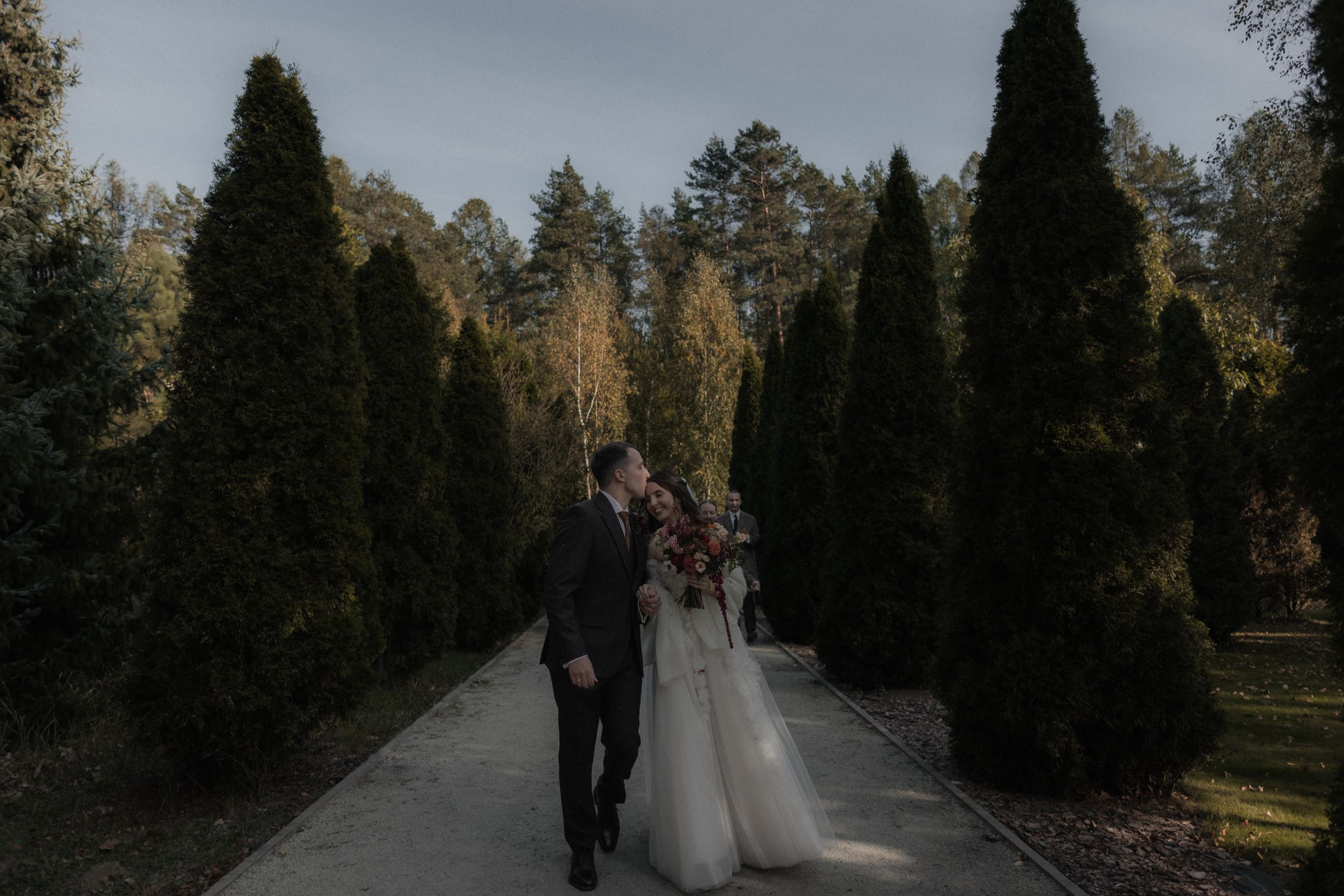 A wedding couple walks along a tree-lined path in a park during autumn with golden sunlight filtering through.