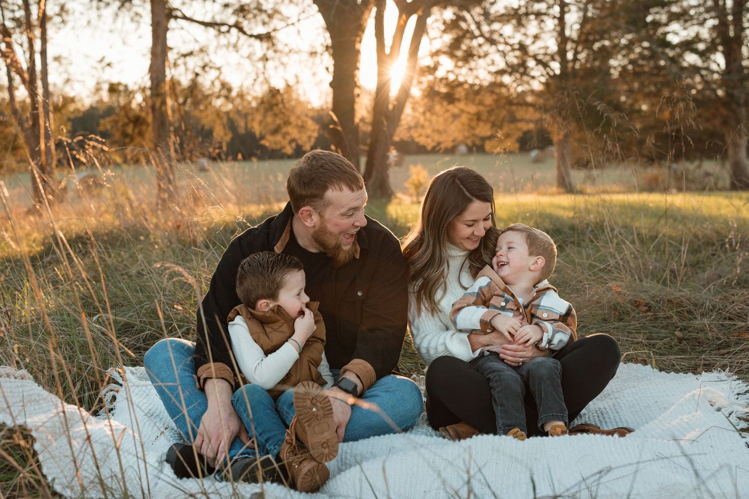 A family plays together in a field during golden hour as sunlight filters through bare winter trees.