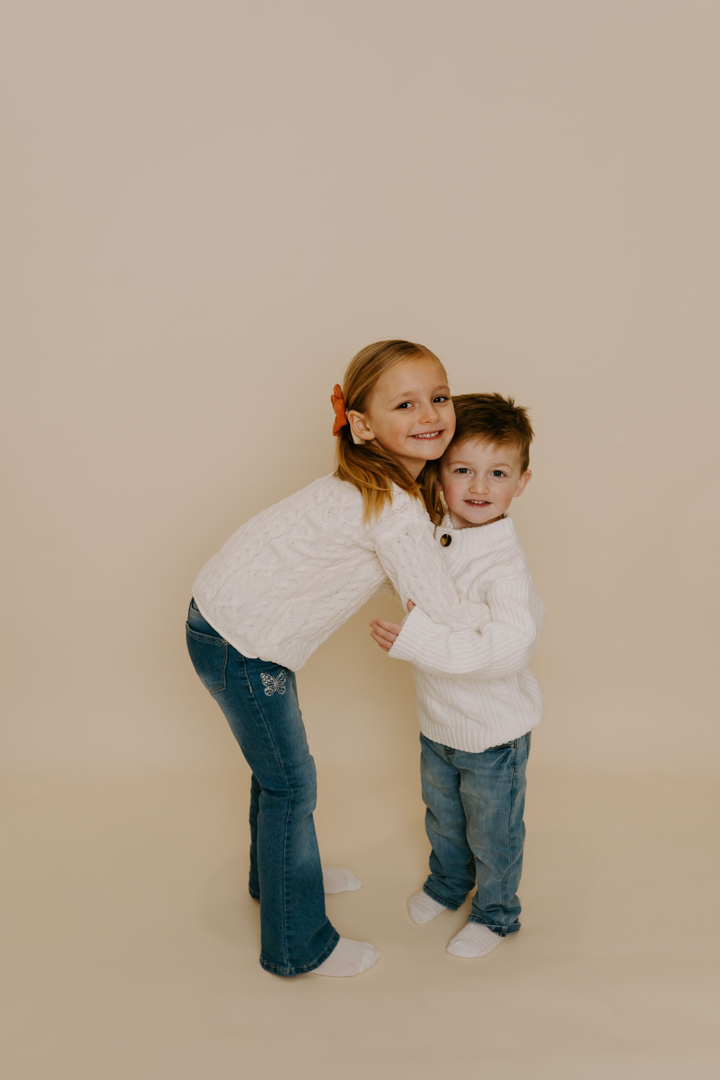 Two children in white sweaters sharing playful moment against neutral background.