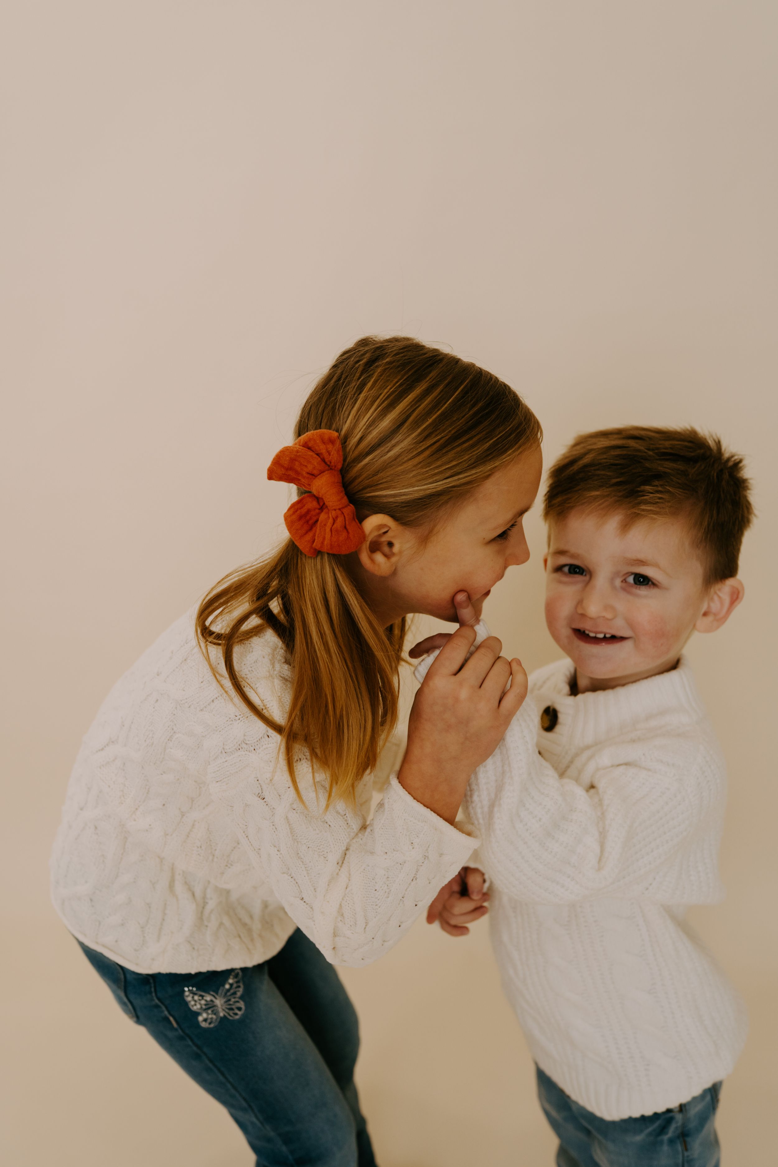 Two children in white sweaters share a playful moment against a beige background.
