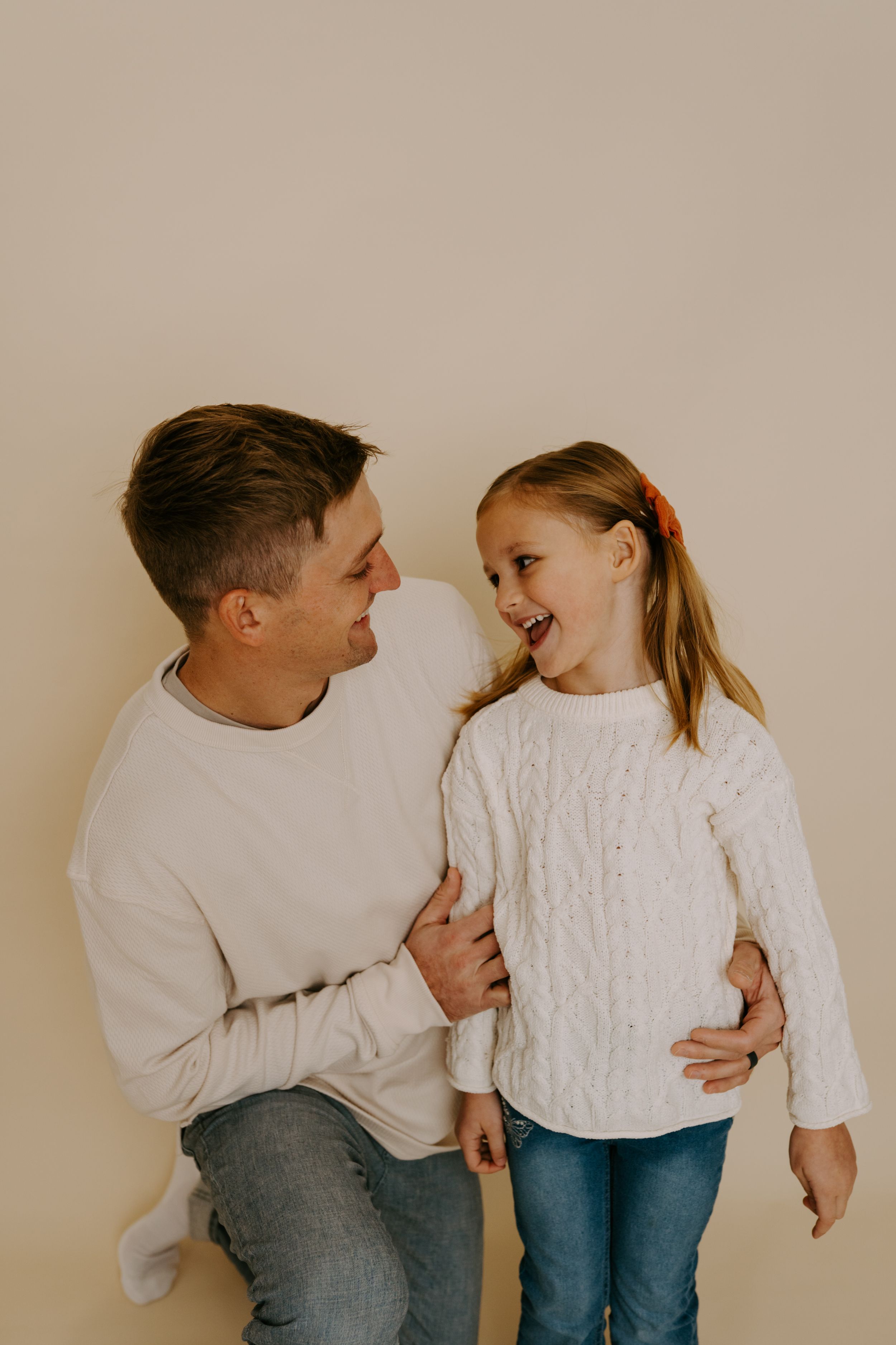 Two people in white sweaters share a joyful laughing moment against beige wall.