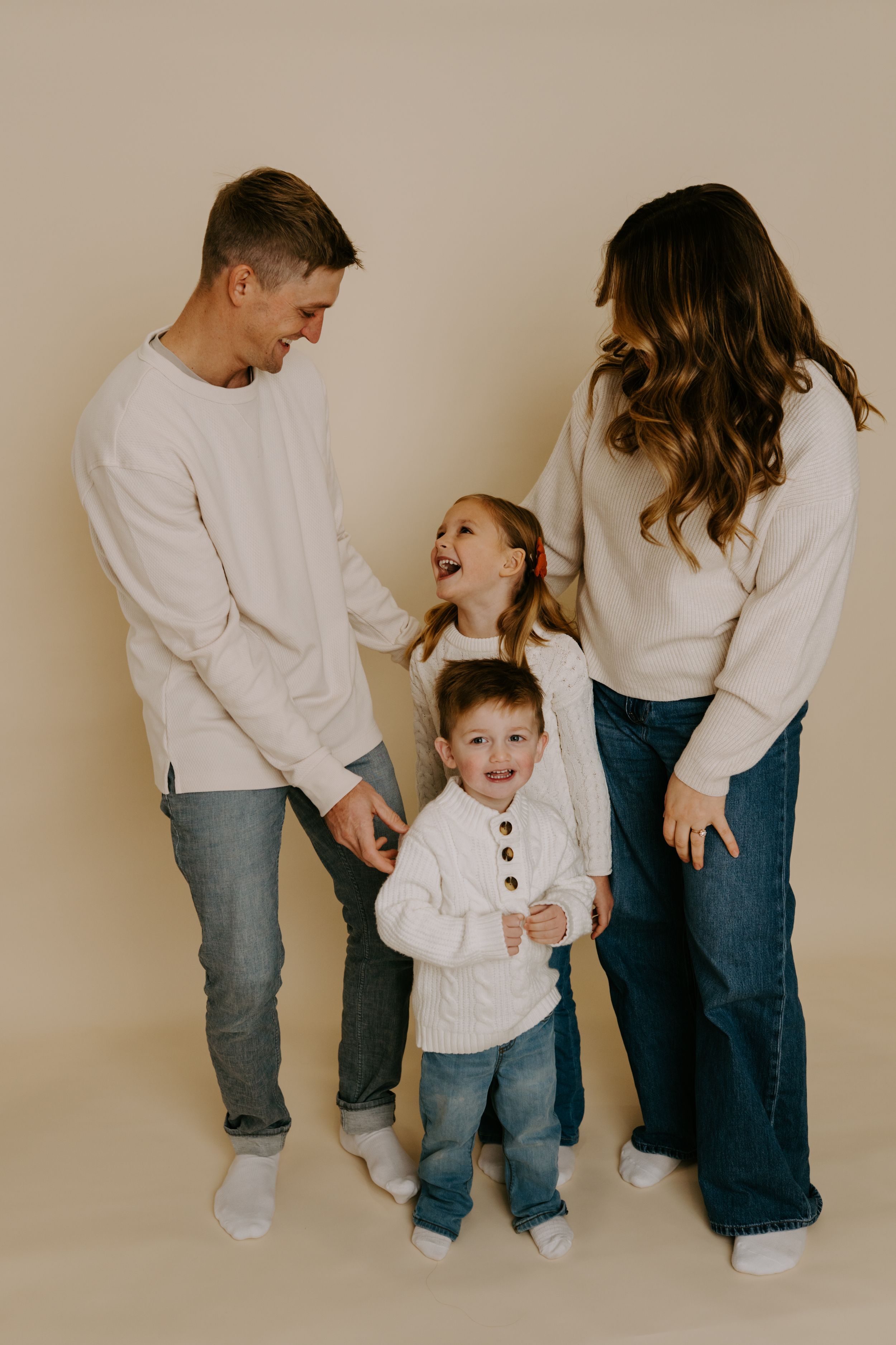 Family of four wearing coordinated white sweaters and jeans share candid moment against neutral background.