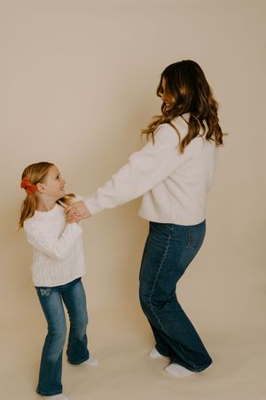 Parent and child in matching white sweaters and jeans dance together against beige background.