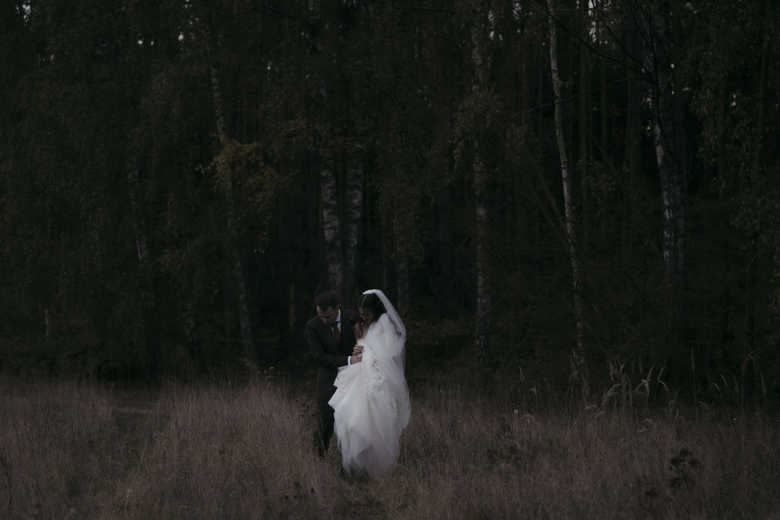 A white chicken walks through tall grass and trees in a dark wooded area.