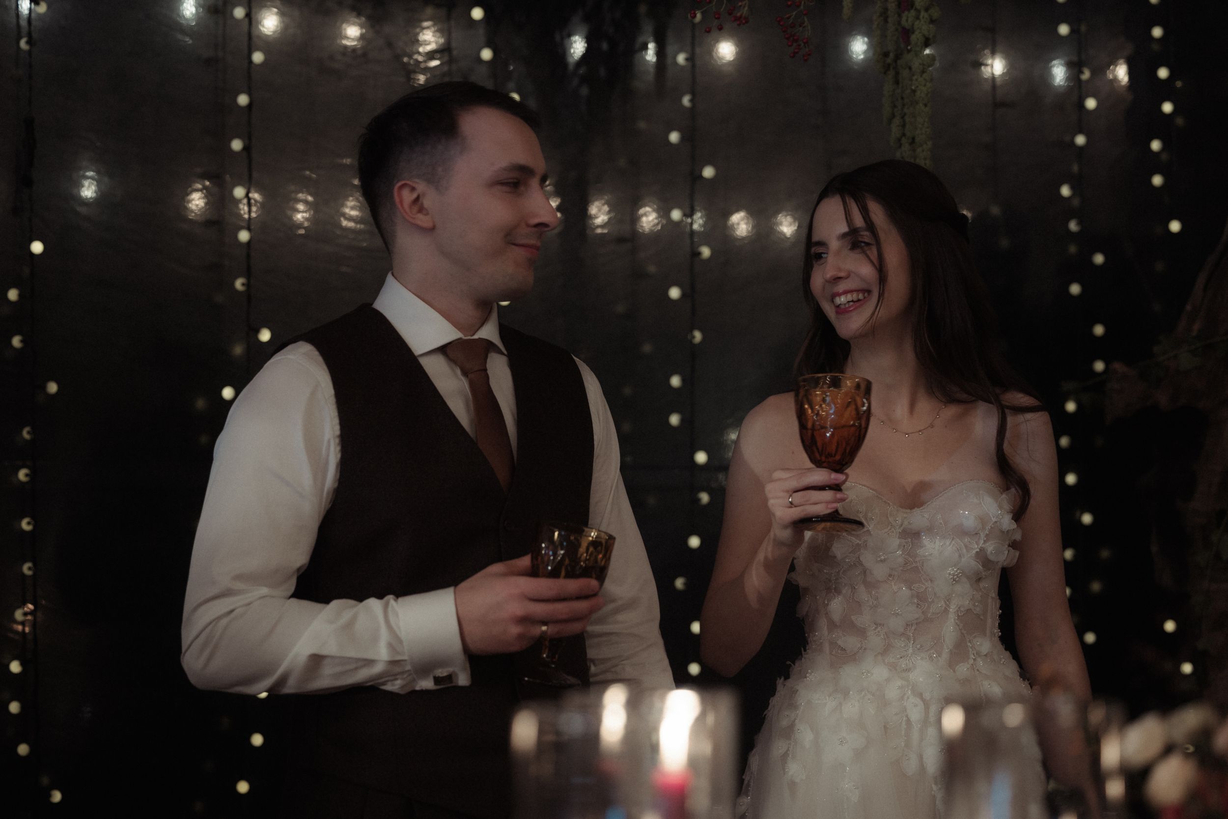 A couple in wedding attire share drinks and laughter during their reception celebration with twinkling lights in background.