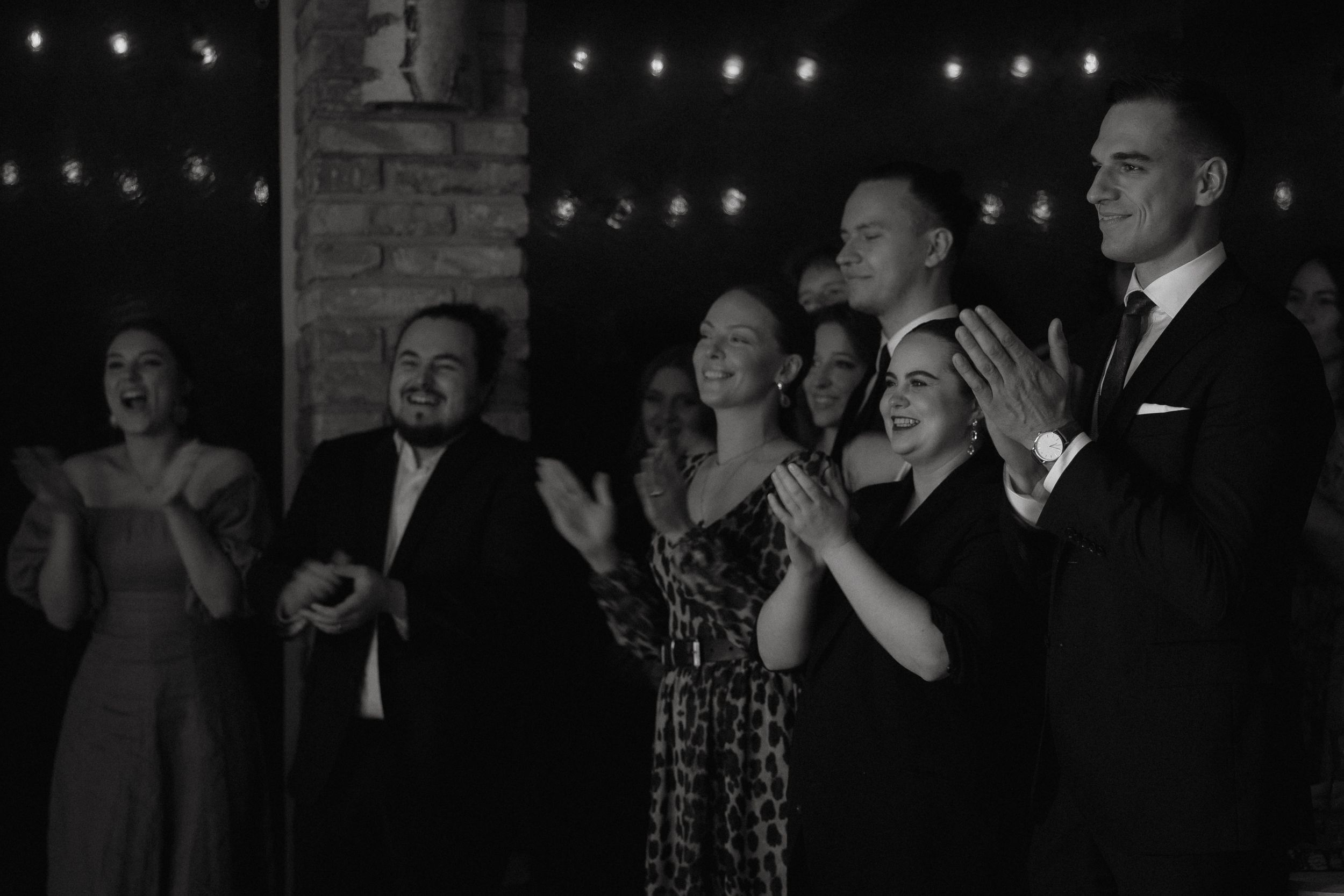 A group of formally dressed people stand together clapping and smiling at a dimly lit event with string lights visible.