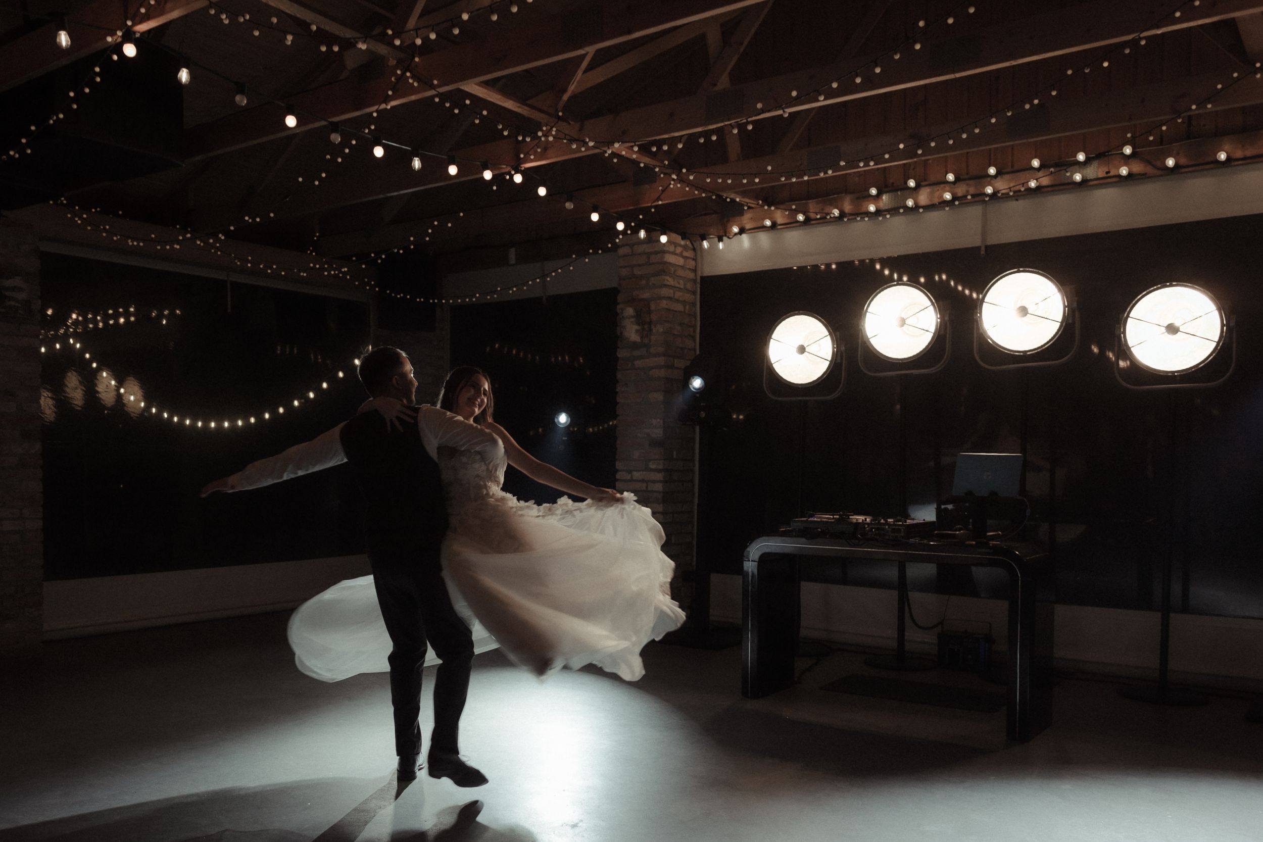 A wedding couple dances under twinkling lights during their first dance at an evening reception.