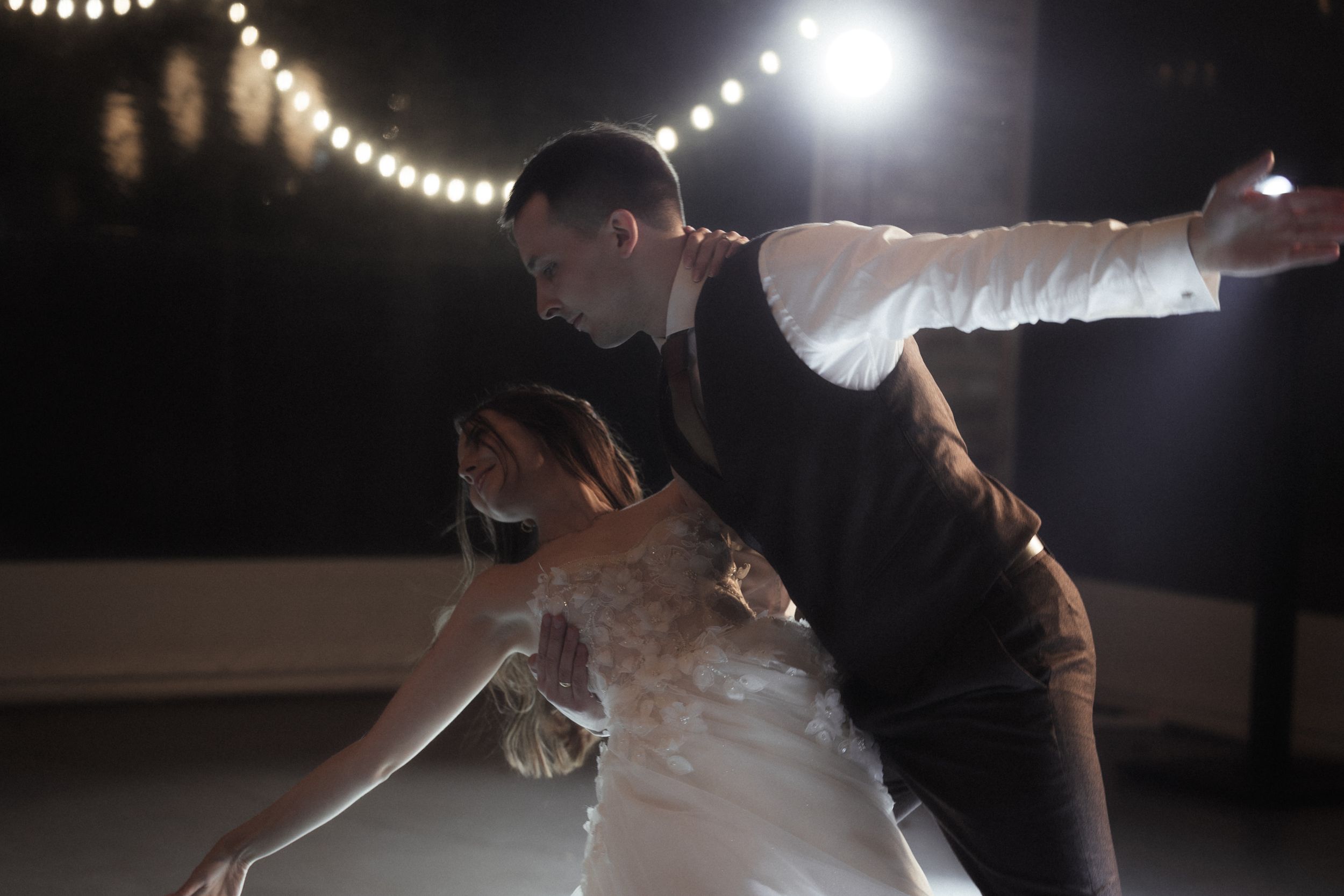A couple performs a romantic dance move during their first dance at a wedding reception under string lights.