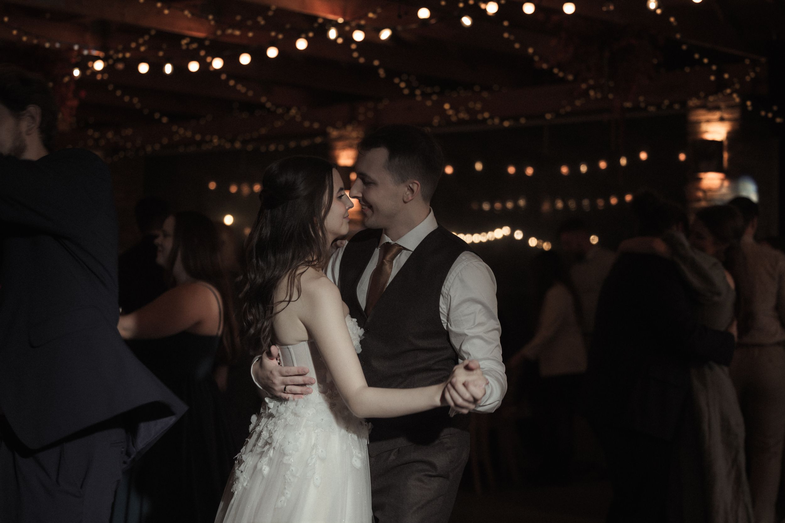 A couple dancing at a wedding reception under warm string lights and dark evening ambiance.