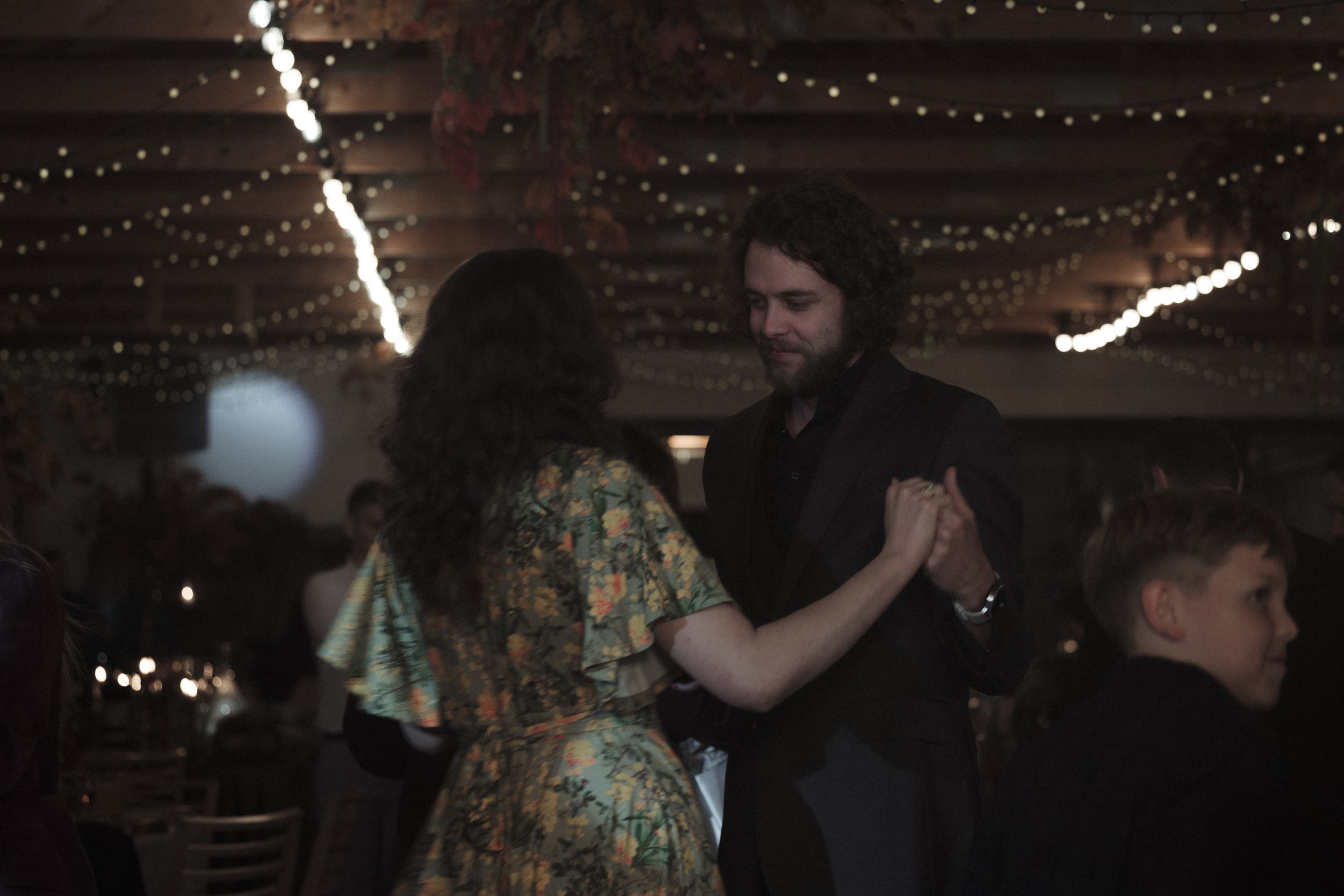 A couple dances together under warm twinkle lights in a dimly lit venue.