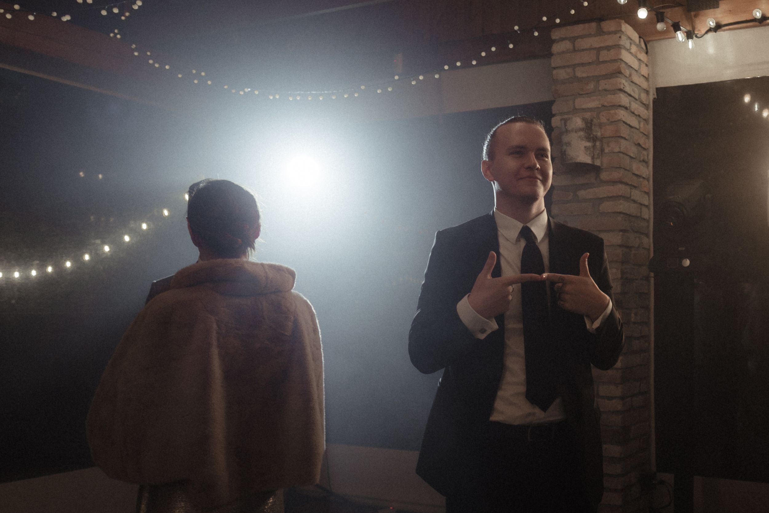 A speaker in a dark room illuminated by string lights gives a presentation at an indoor event.