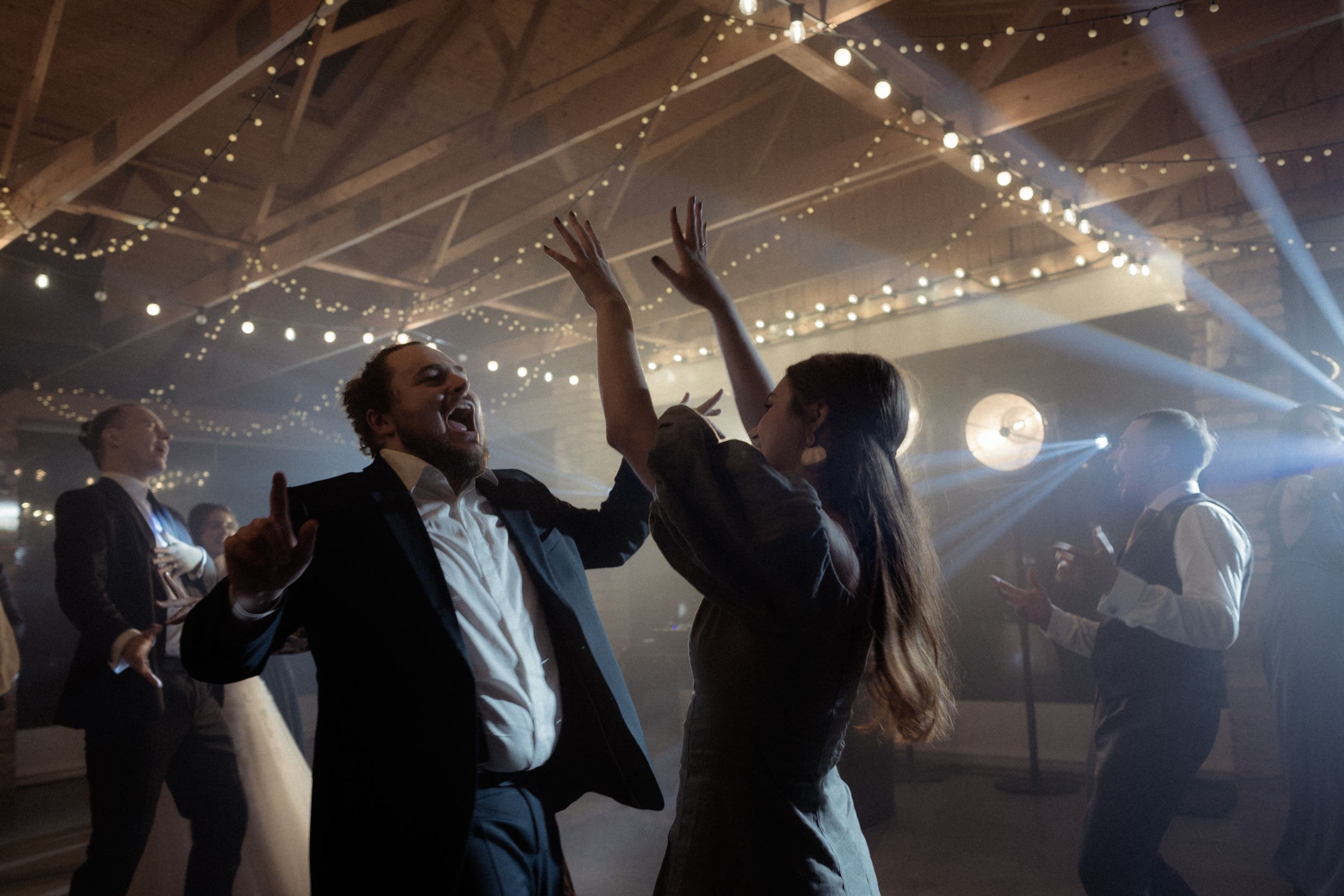 People dancing and celebrating at a wedding reception under string lights and atmospheric lighting.