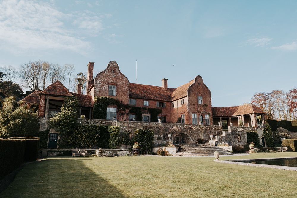 Historic brick manor house with multiple wings and chimneys stands on a green lawn under blue skies.