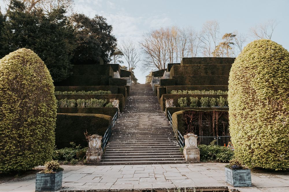 Stone steps lead upward through a formal garden with neatly trimmed hedges and ornamental statues on a cloudy day.
