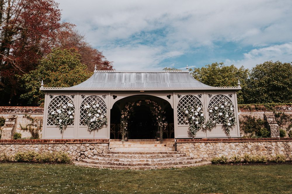 A white latticed garden pavilion with stone walls and arched entrance sits in a landscaped garden setting.