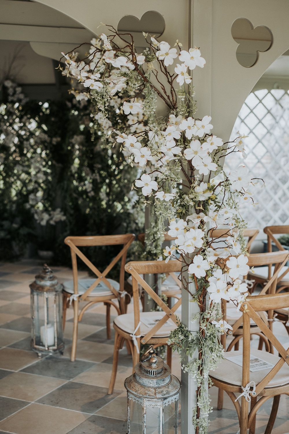 Elegant wedding venue setup with wooden crossback chairs and tall white floral arrangements on stone flooring.