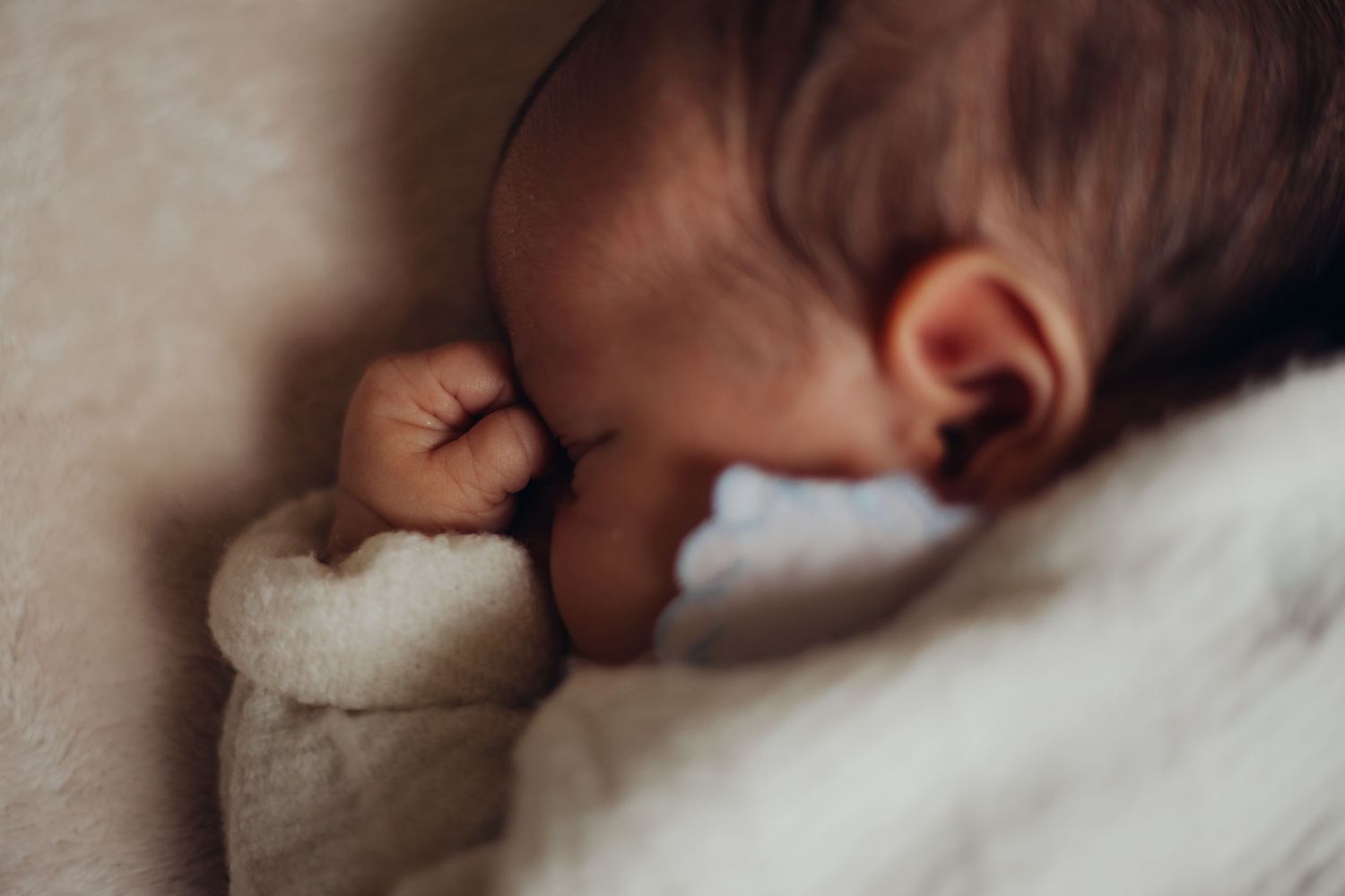 Sleeping newborn baby with tiny fist clenched, resting peacefully on a soft white blanket.