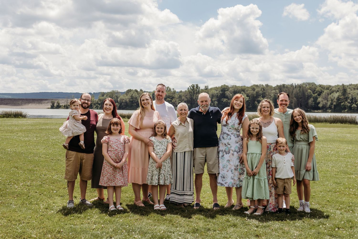 Group photo of wedding party in pastel dresses and suits standing together in a sunny outdoor field setting.