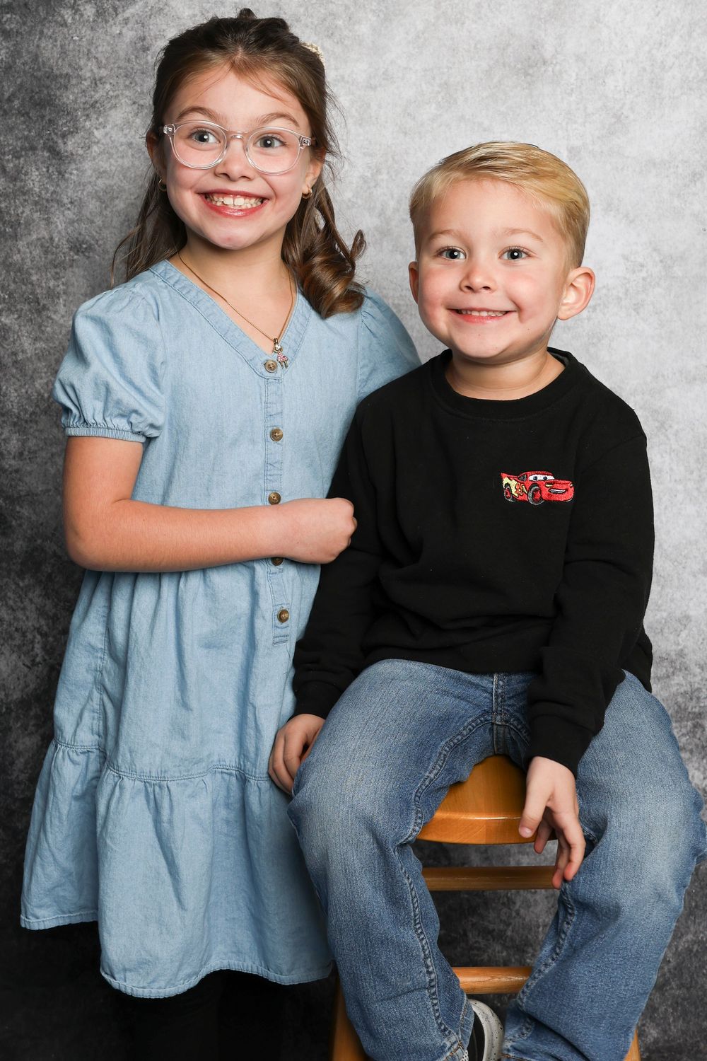 Siblings sitting together on wooden stool wearing denim and casual attire.