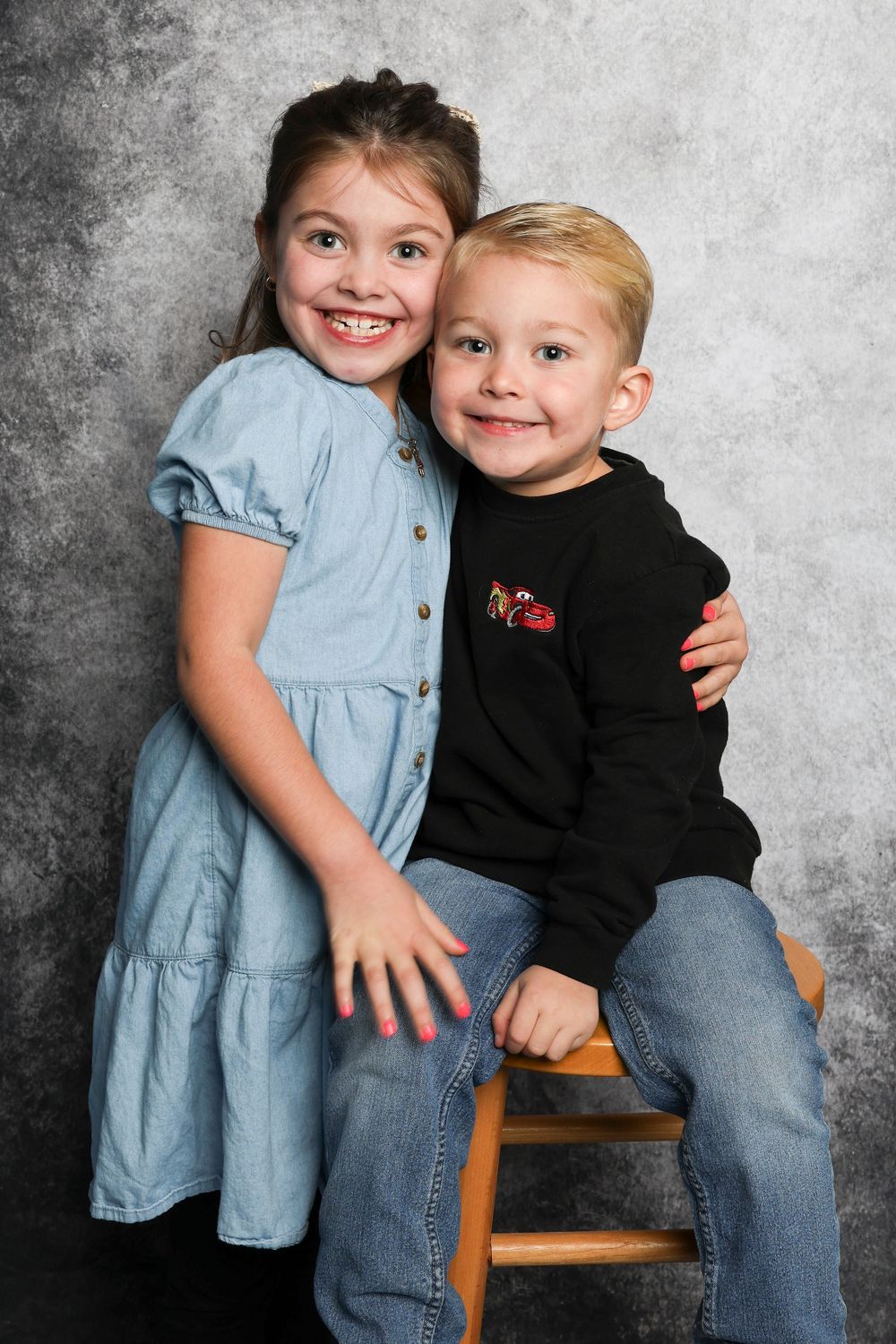 Two children posing together on wooden stool, wearing coordinating blue dress and black sweater.