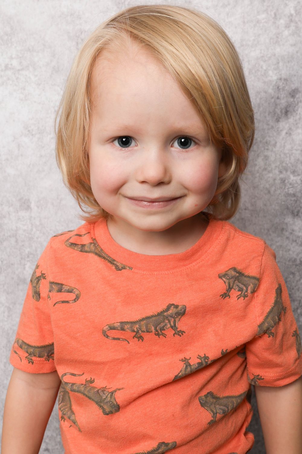 Child wearing bright orange t-shirt with dinosaur prints against grey background.