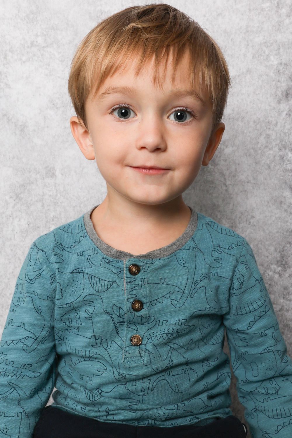 Child wearing a teal shirt with dinosaur patterns against a gray background.