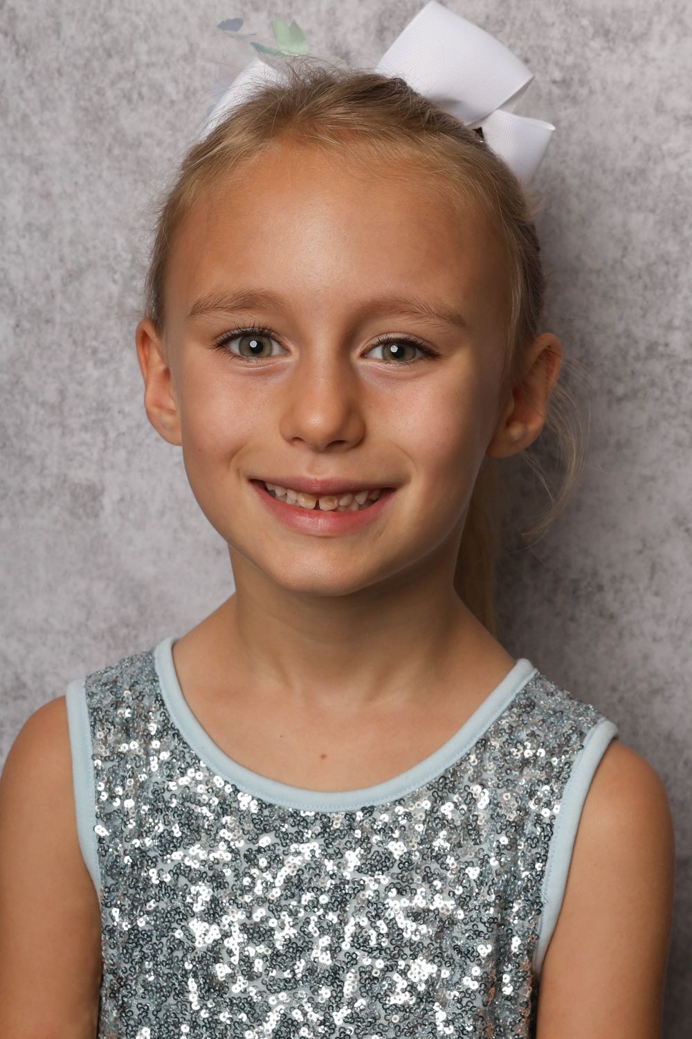 Young student wearing a sparkly silver sequined tank top and white bow against a gray background.
