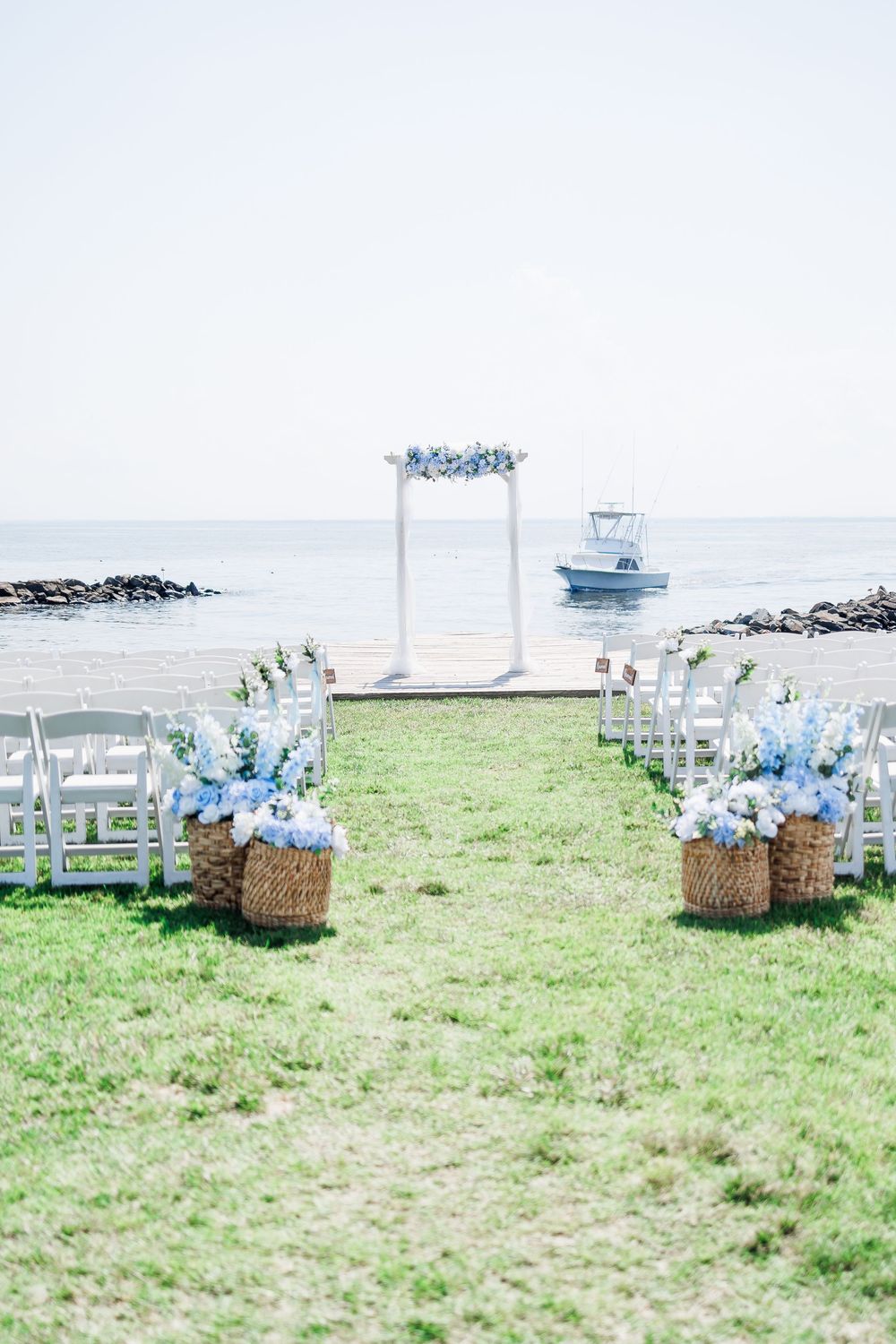 White chairs arranged on green grass for an outdoor waterfront wedding ceremony with blue floral decorations.