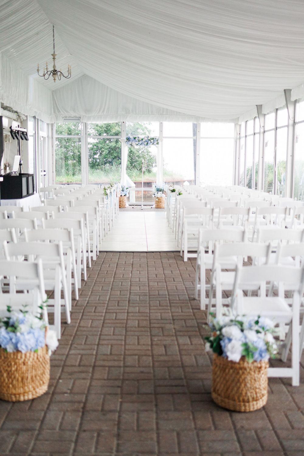 White wedding ceremony setup with draped tent ceiling, rows of chairs, and blue floral arrangements in wicker baskets.