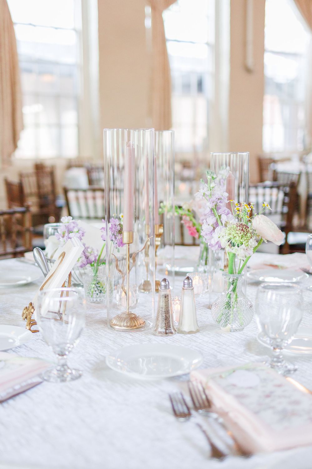 Elegant wedding reception tables set with white linens, pink floral centerpieces, and crystal glassware in a bright venue.