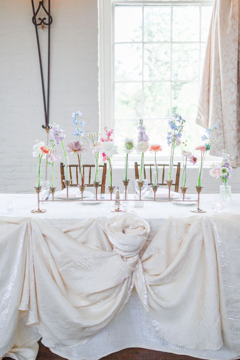 Long white table decorated with delicate floral arrangements in glass vases set against bright windows in an elegant room.