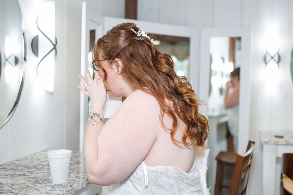 A bride in a white gown adjusts her dress while getting ready in a brightly lit room with mirrors.
